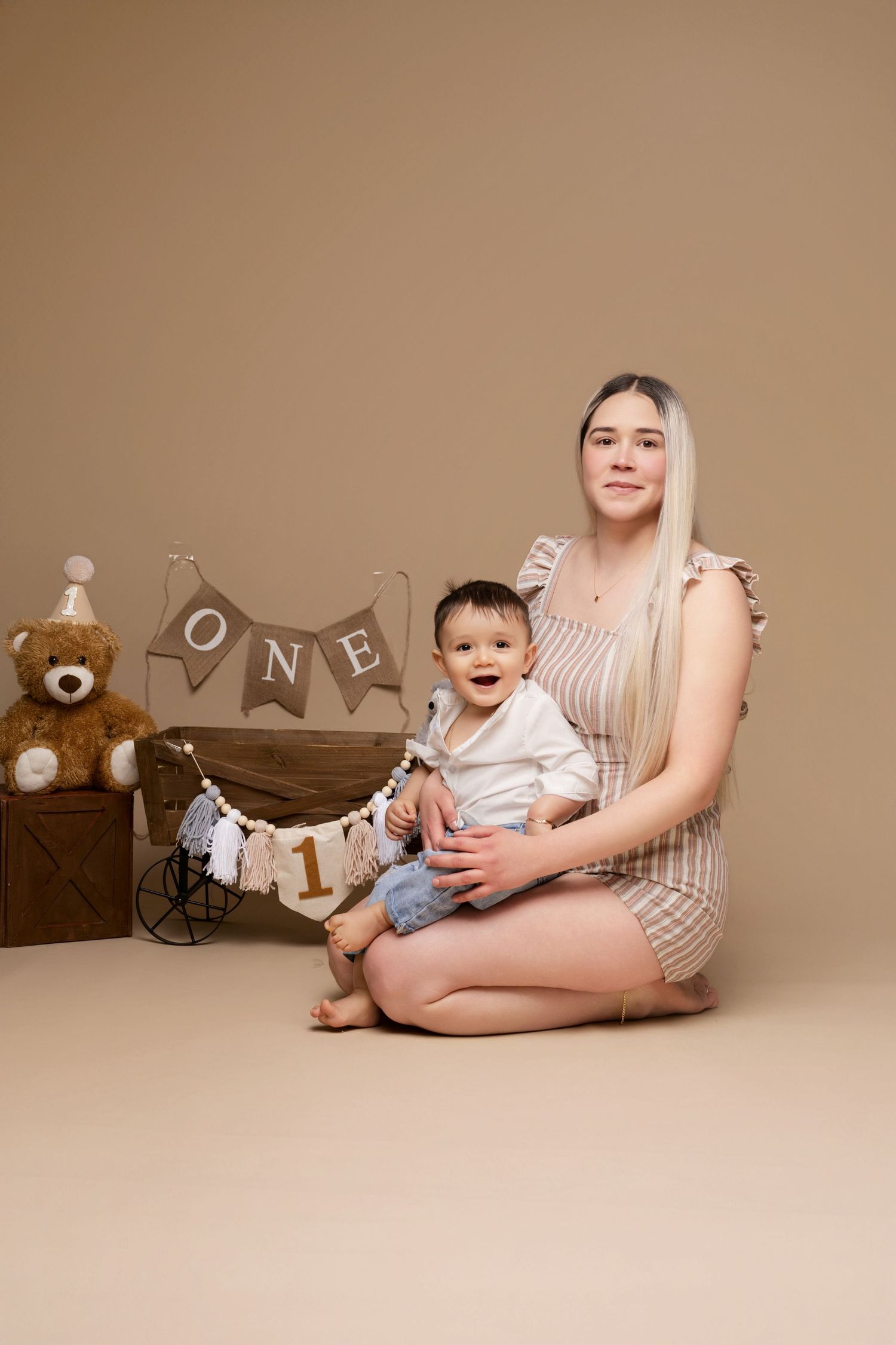 A young mother sits cross-legged holding her smiling baby at a first birthday photo shoot with ONE banner and teddy bear.