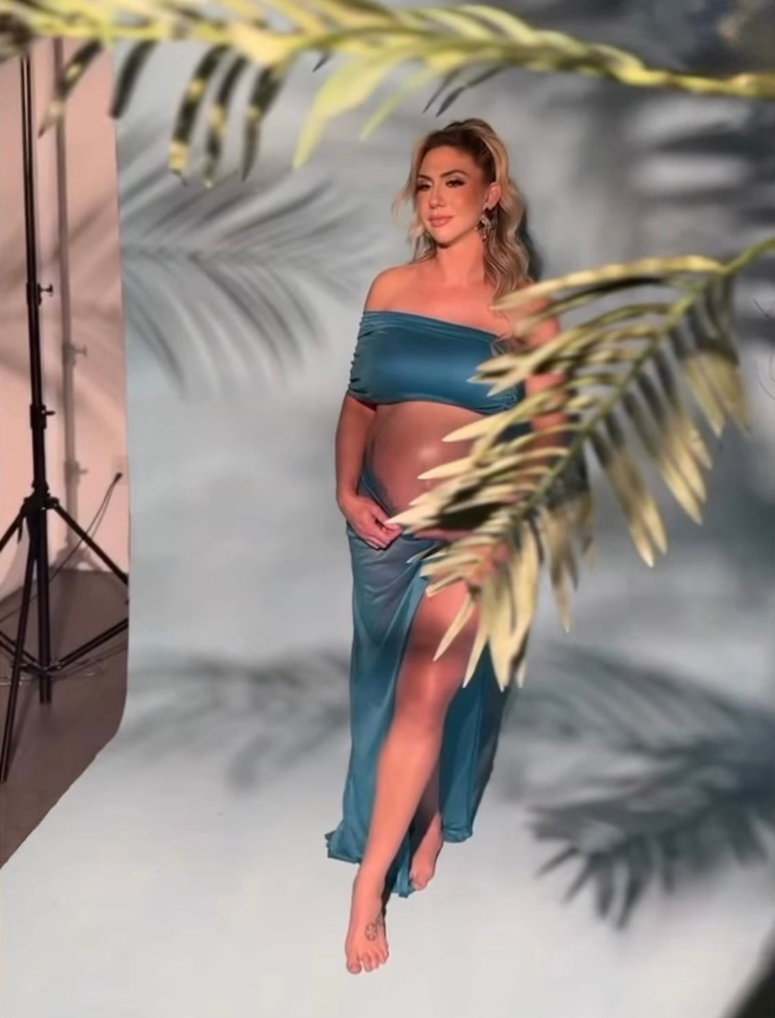 Pregnant woman in teal off-shoulder crop top and slit maxi skirt posing barefoot in tropical photo studio.