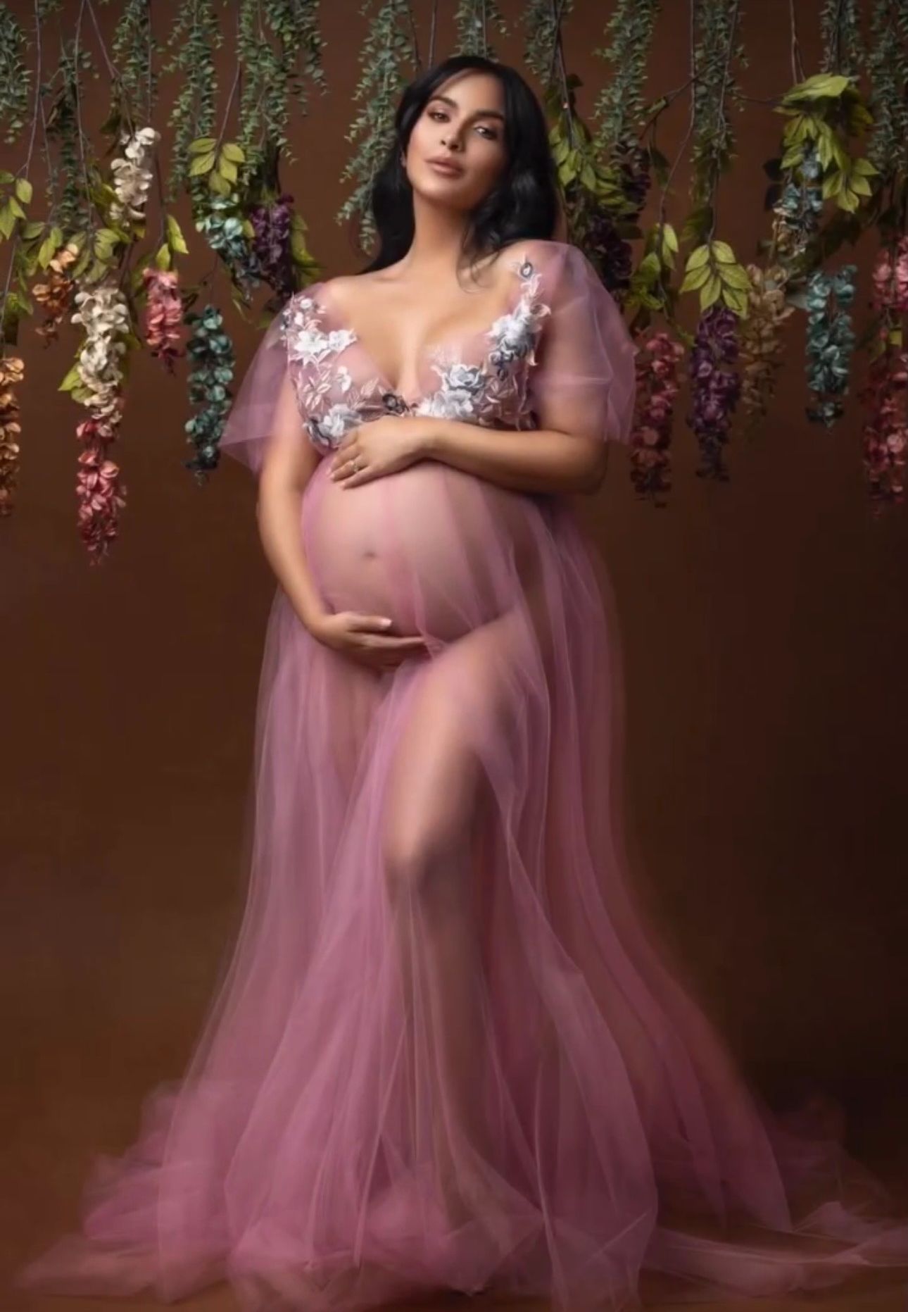 Pregnant woman in sheer pink floral tulle maternity gown poses gracefully with hanging flowers backdrop.