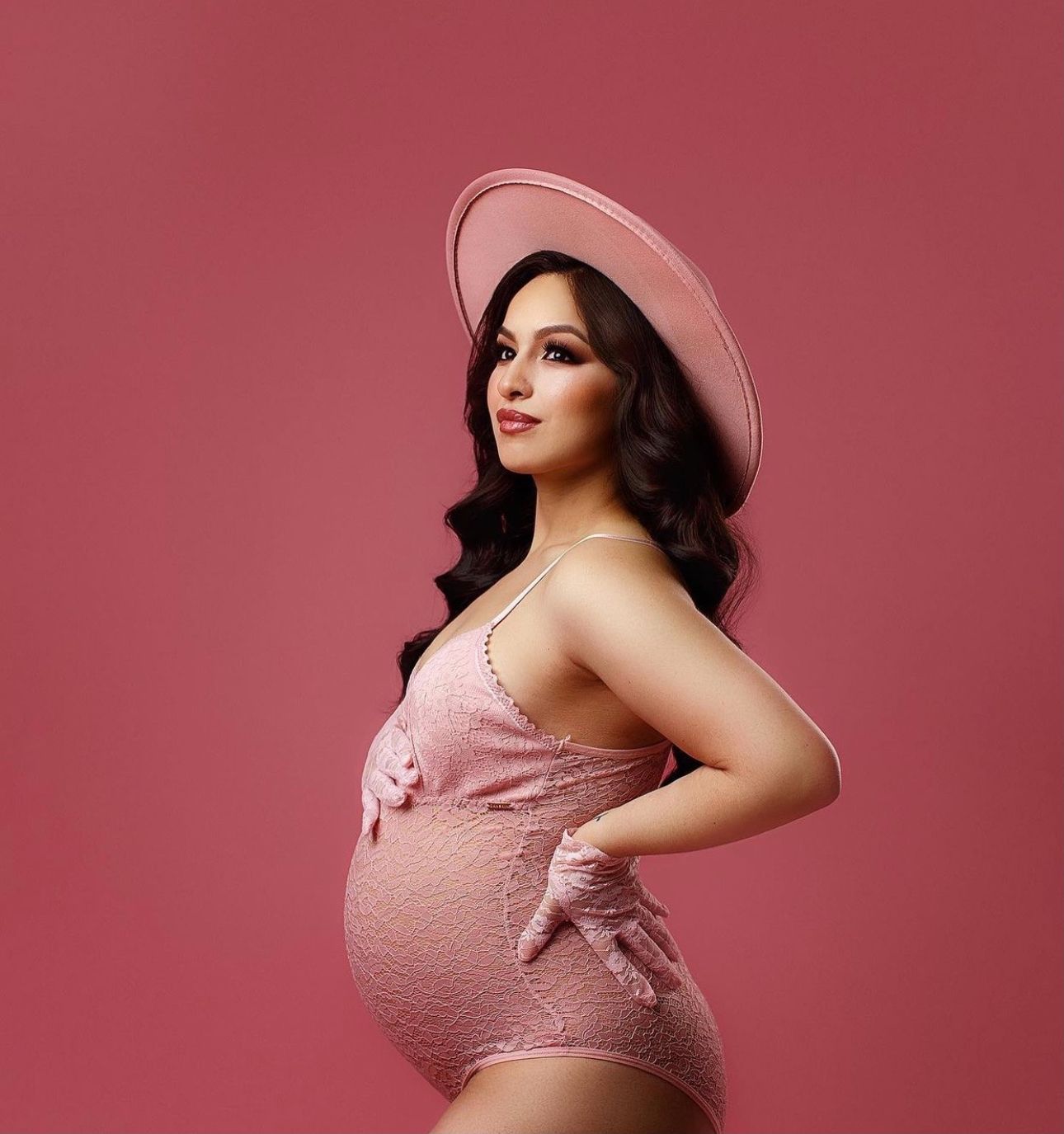 Pregnant woman in pink lace bodysuit and wide-brim hat posing gracefully against a pink background.