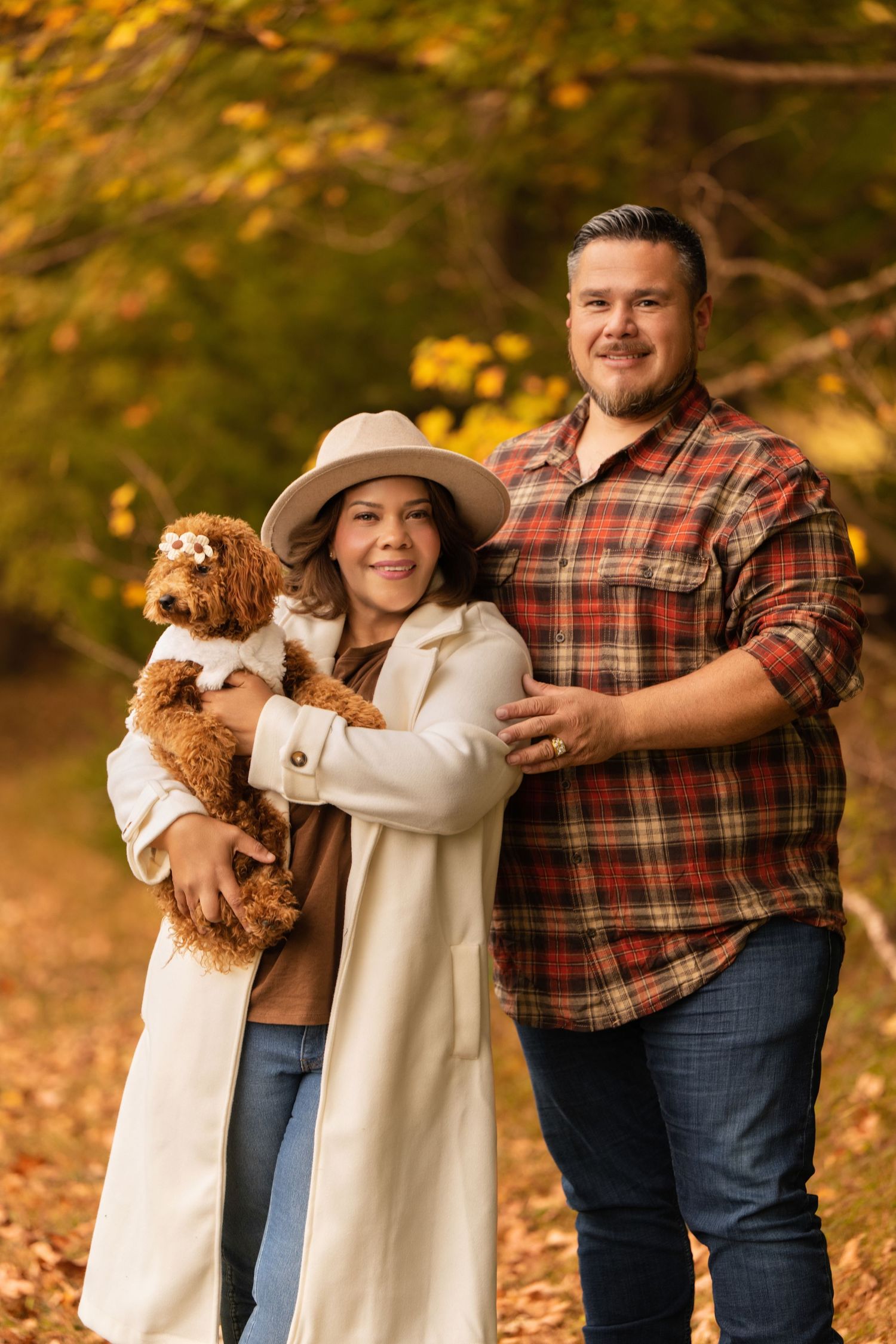 Smiling couple holding a fluffy brown dog in an autumn forest setting, woman wearing a beige hat and white coat.