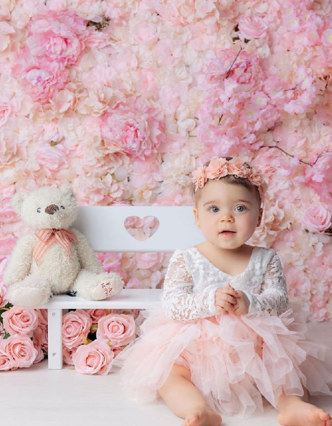 A baby in a pink tutu dress poses against a romantic floral backdrop of pink roses and blossoms.