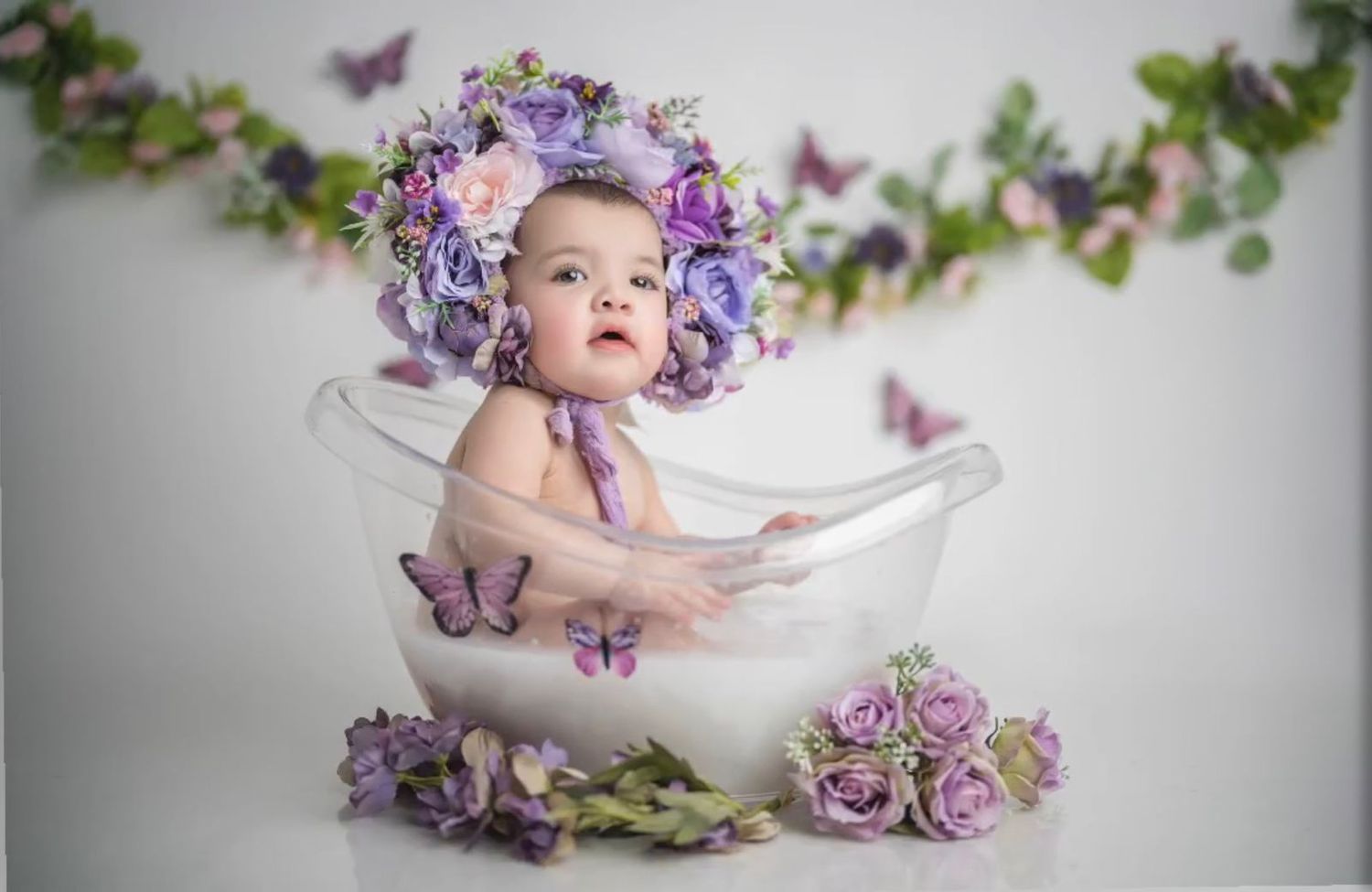 Baby in bathtub photo session with purple flowers and butterfly decorations against white backdrop.