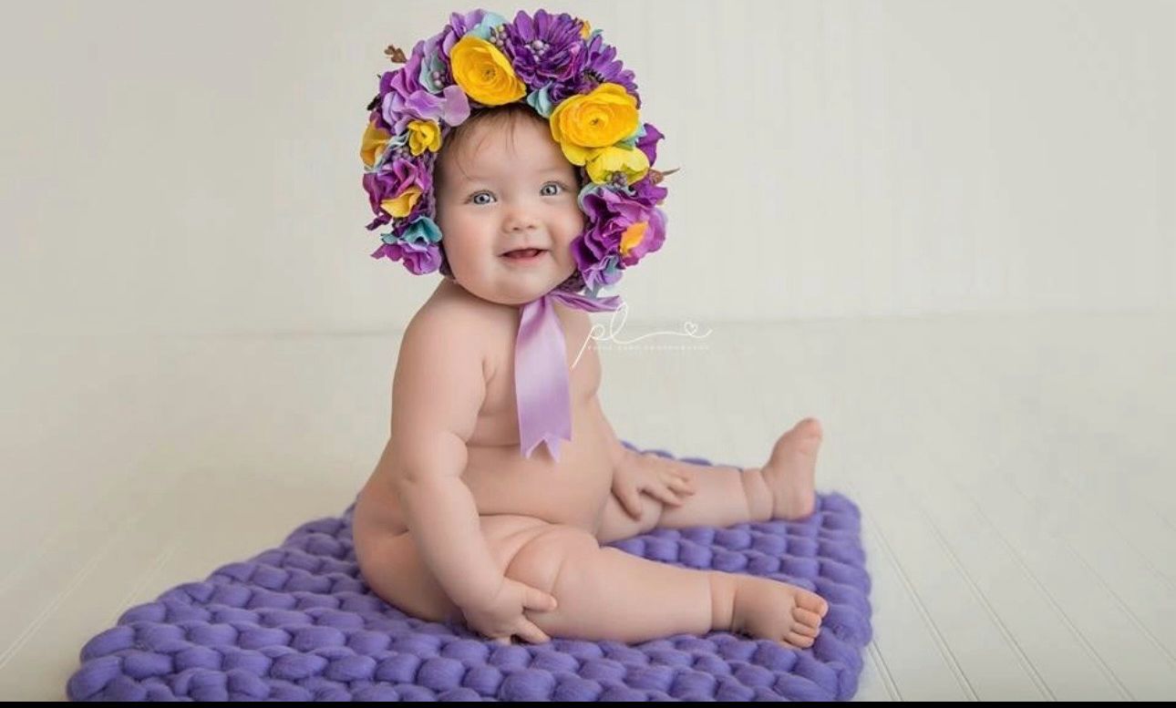 Baby wearing vibrant purple and yellow floral crown sitting on purple knit blanket.