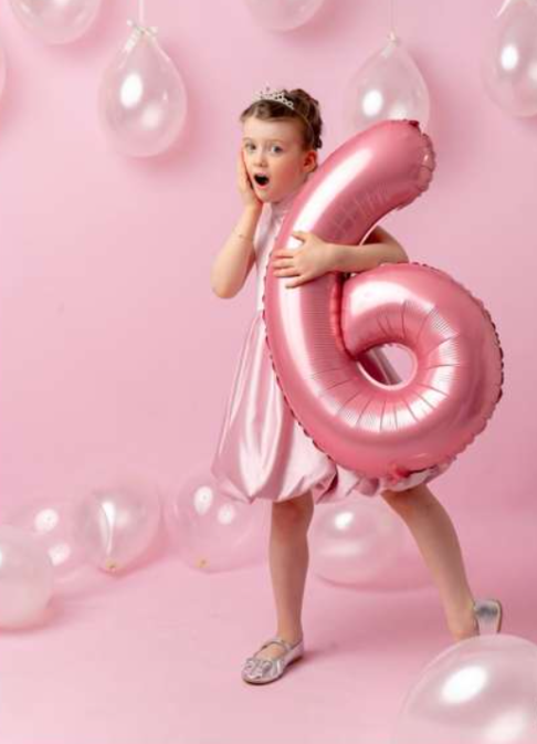 A surprised little girl in a pink dress holds a large pink number 6 balloon, surrounded by white balloons on a pink background.