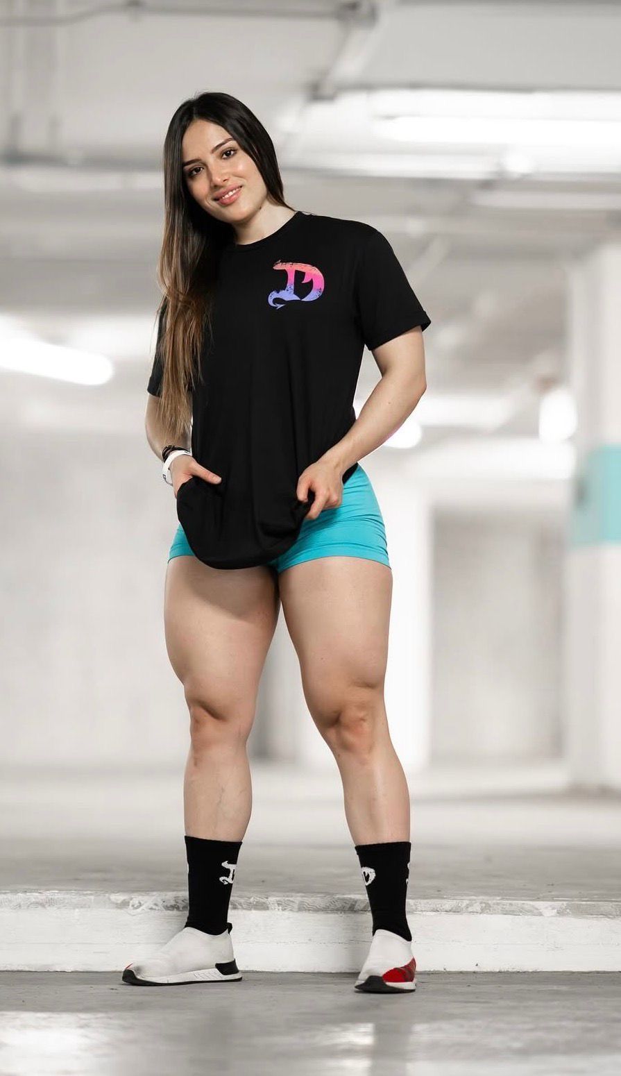 Fit woman in black graphic tee, teal shorts, and white sneakers posing in a parking garage.