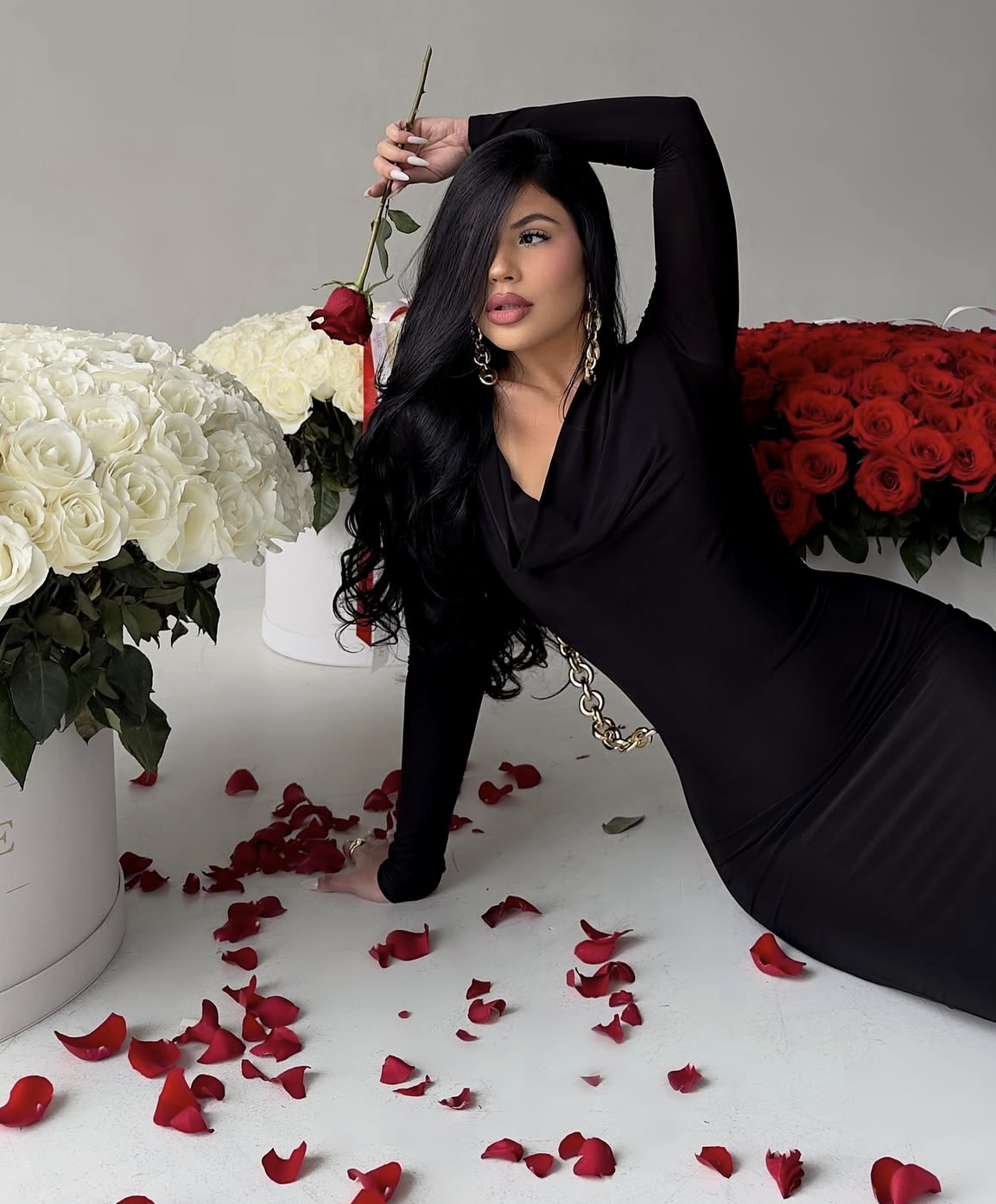 Woman in black dress poses with red rose among white and red rose bouquets with scattered petals on floor.