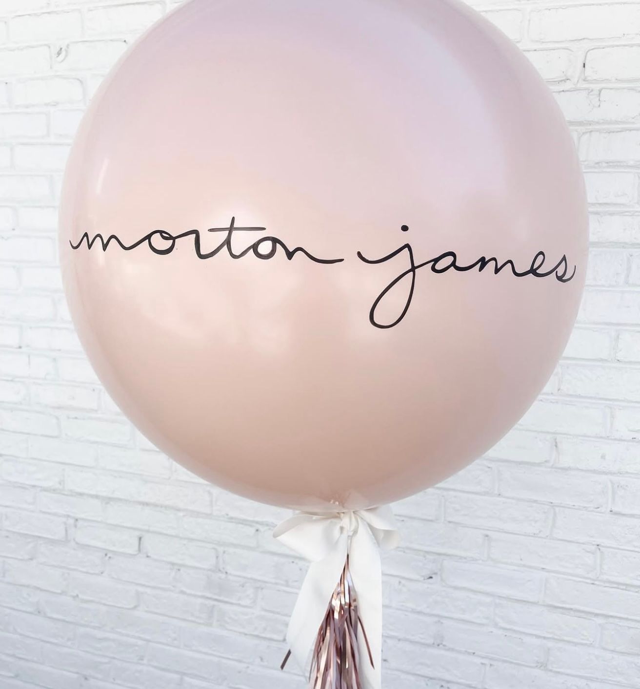Large blush pink balloon with elegant black cursive script reading morton james, adorned with white ribbon and rose gold tassel.