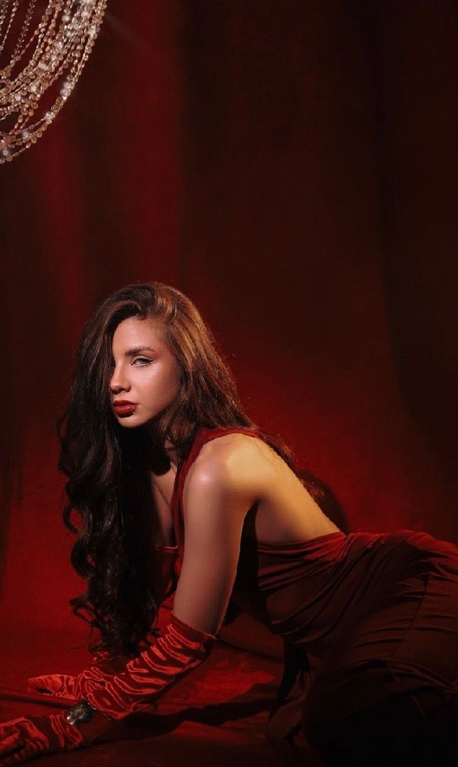 Elegant woman in red gown with long gloves posing dramatically against a rich red backdrop with crystal chandelier.