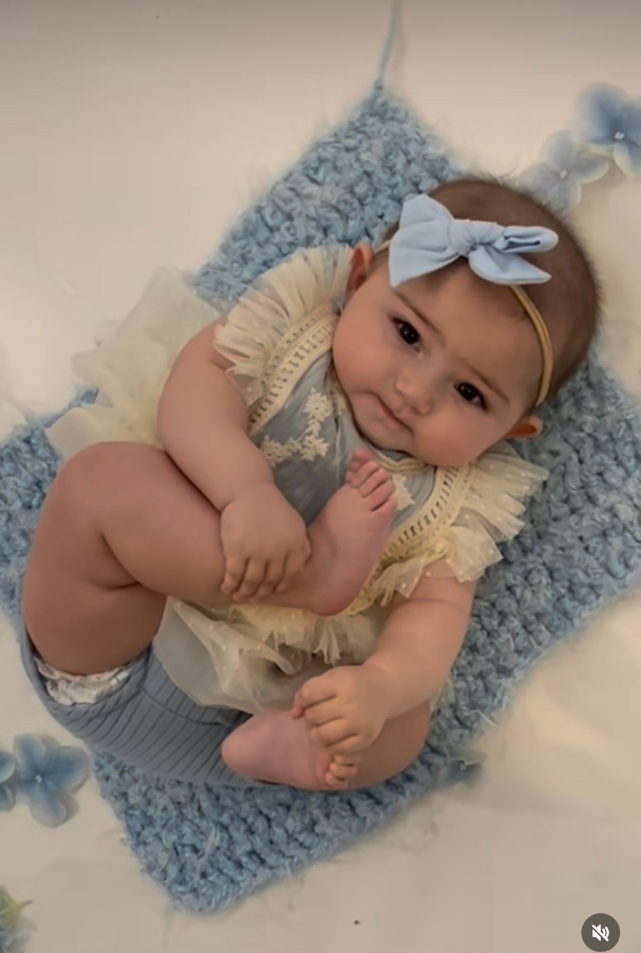 Adorable baby girl in cream ruffle romper and blue bow headband lying on blue knit blanket.