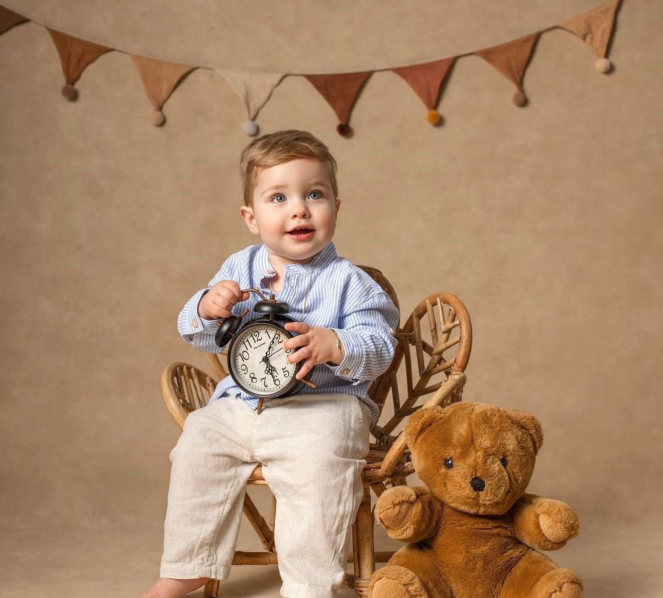 A cute toddler boy sits on a wooden chair holding a clock beside a teddy bear with bunting flags in background.