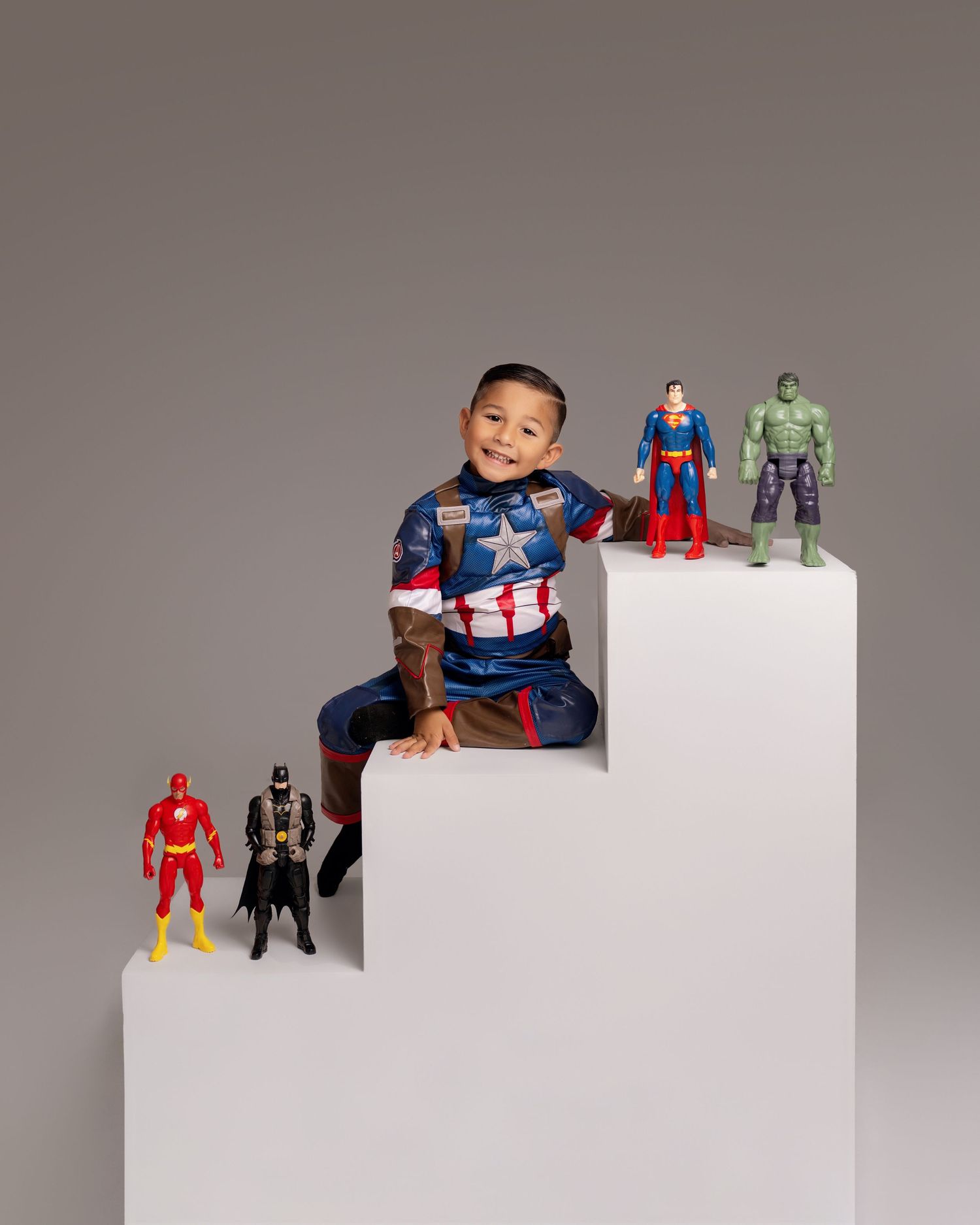 Boy in Captain America costume sits on white steps with Superman, Hulk, Batman, and Flash action figures.