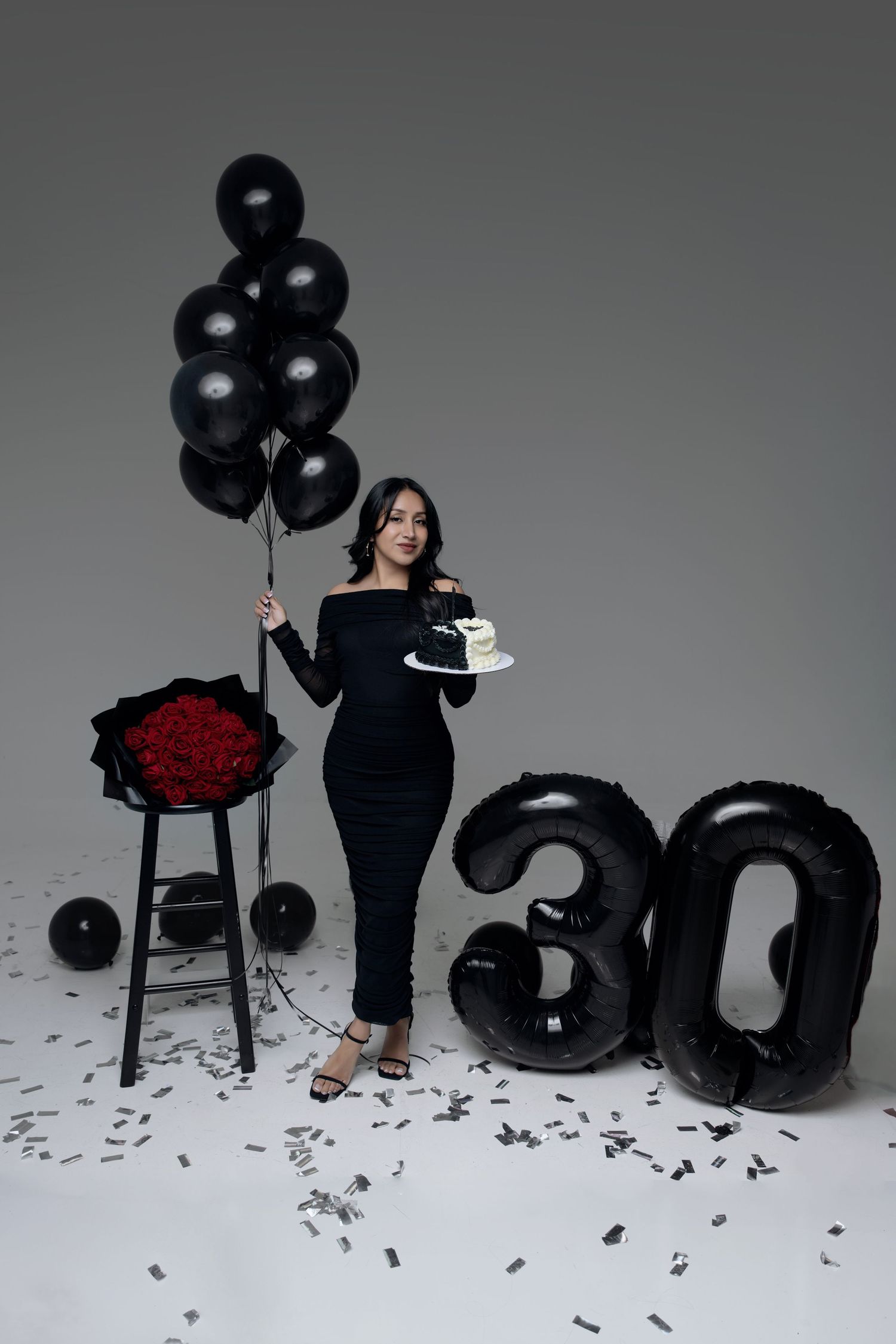 Woman in black dress holding balloons and cake beside red roses and number 30 balloons for a birthday celebration.