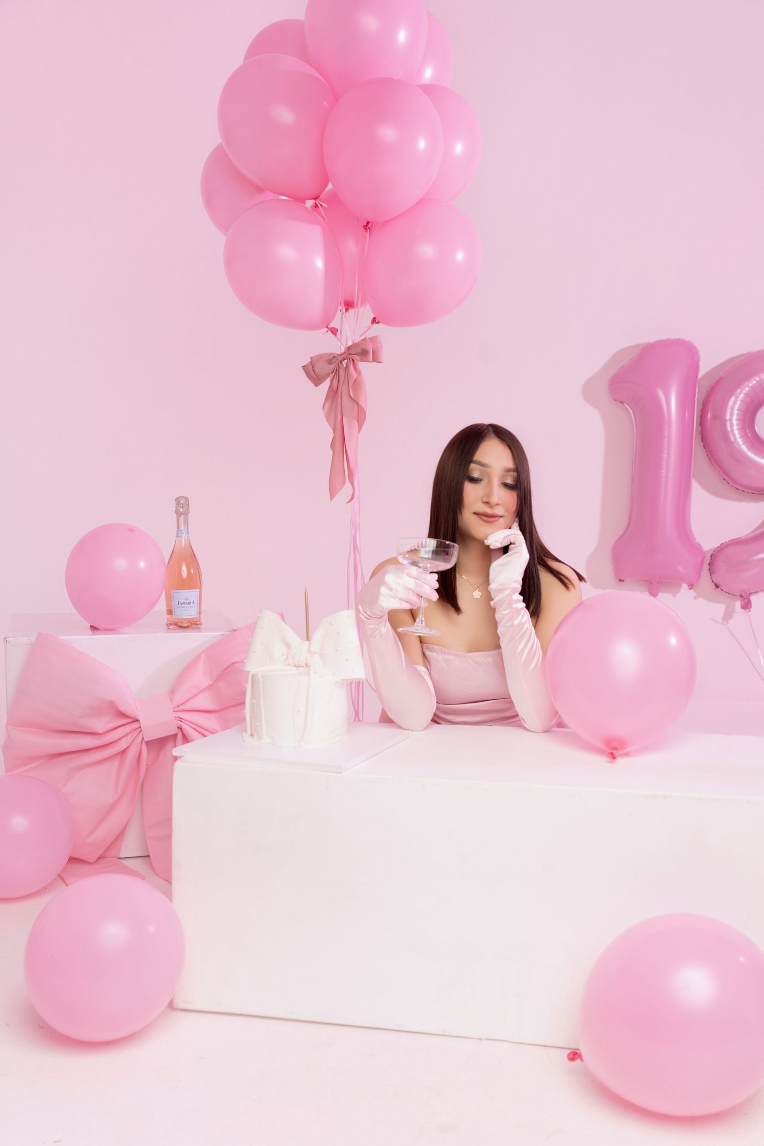 Young woman in pink outfit celebrating 18th birthday with balloons, gifts, and champagne in pink decorated studio.