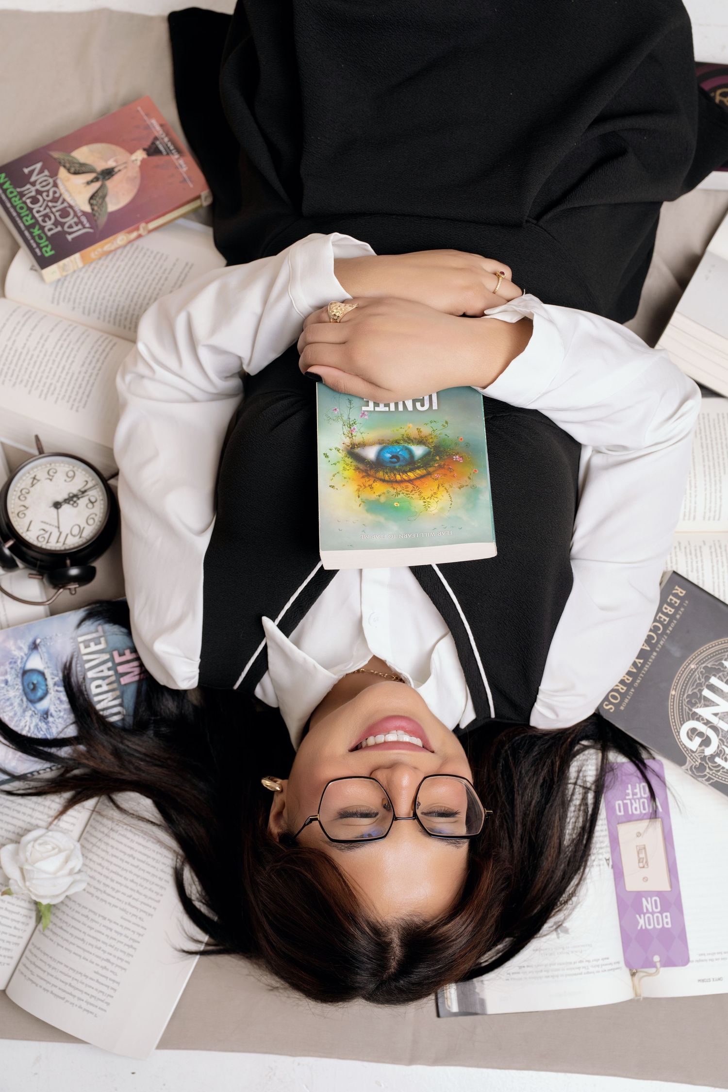 Smiling girl with glasses lies on her back among open books, holding a colorful novel with an eye on the cover beside a vintage alarm clock.