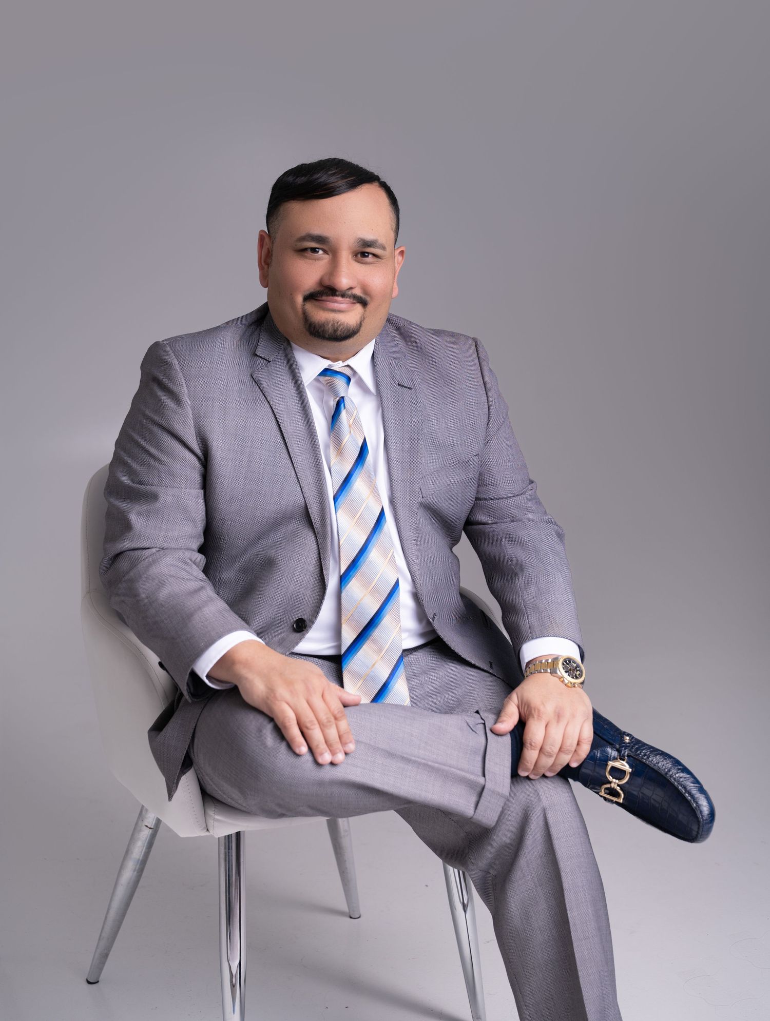 Smiling man in a gray suit and blue striped tie sits crossed-legged on a white modern chair against a neutral gray studio backdrop.