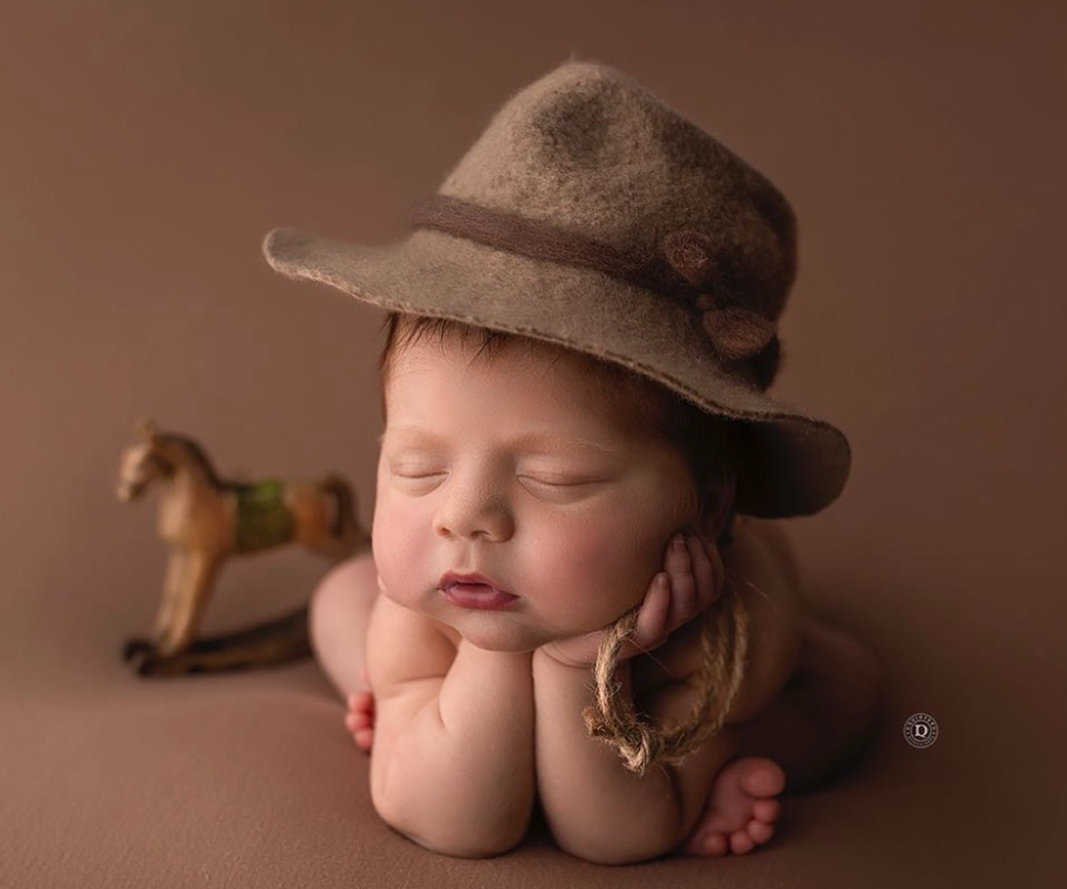 A sleeping baby wearing a brown newsboy cap lies peacefully on a brown backdrop in a professional portrait setting.