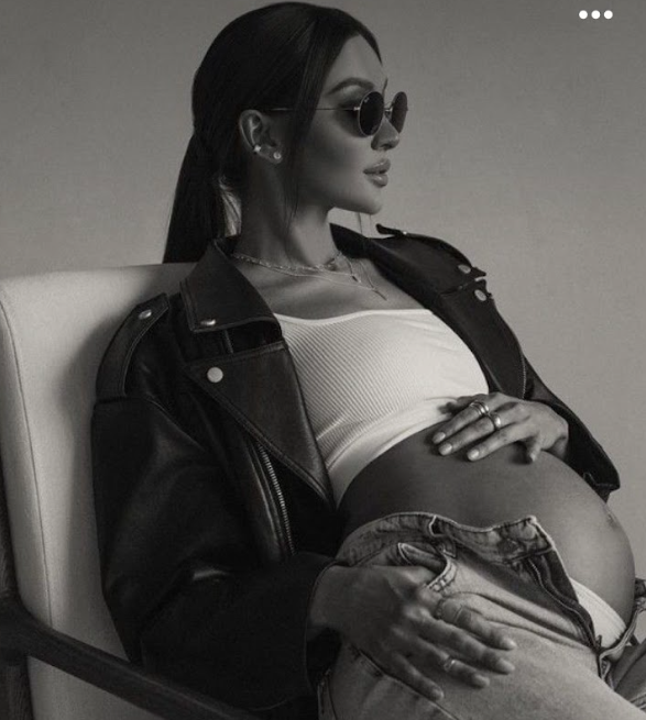 Black and white photo of a pregnant woman in sunglasses and leather jacket sitting in a chair.