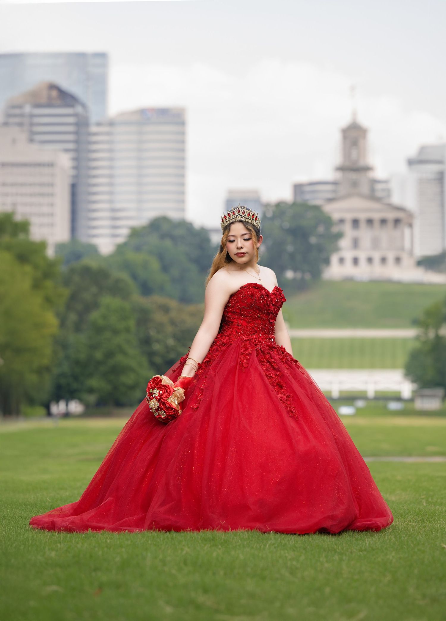 Young woman in red ball gown and crown holds bouquet on green lawn with city skyline backdrop.