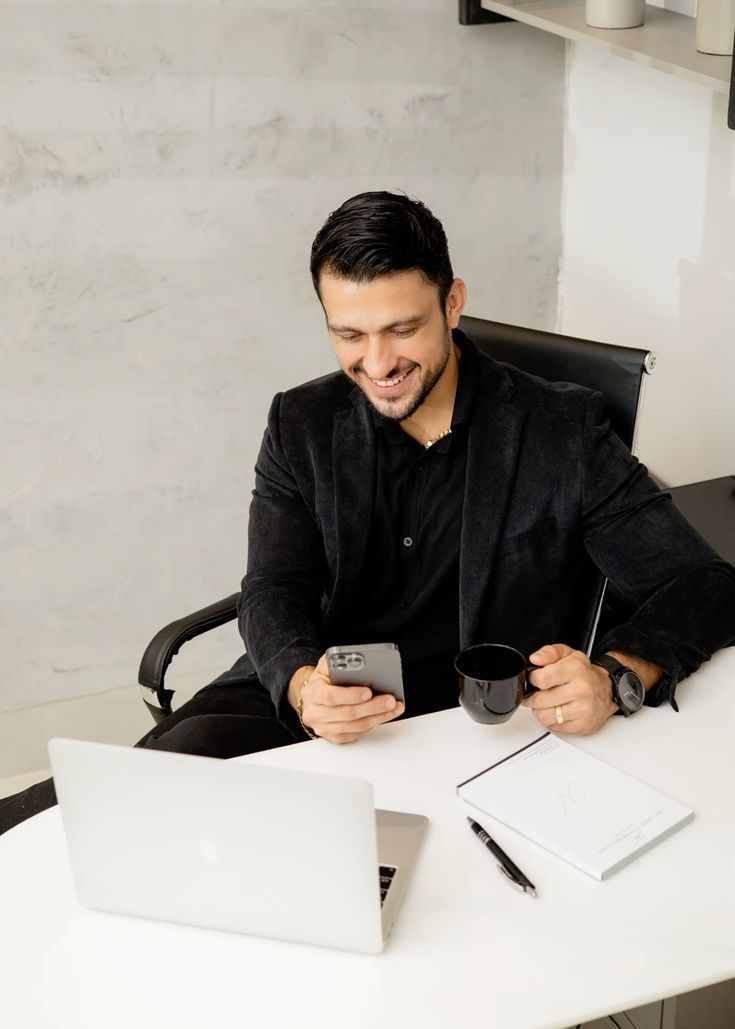 Smiling businessman in black suit using smartphone at desk with laptop and coffee cup in modern office.