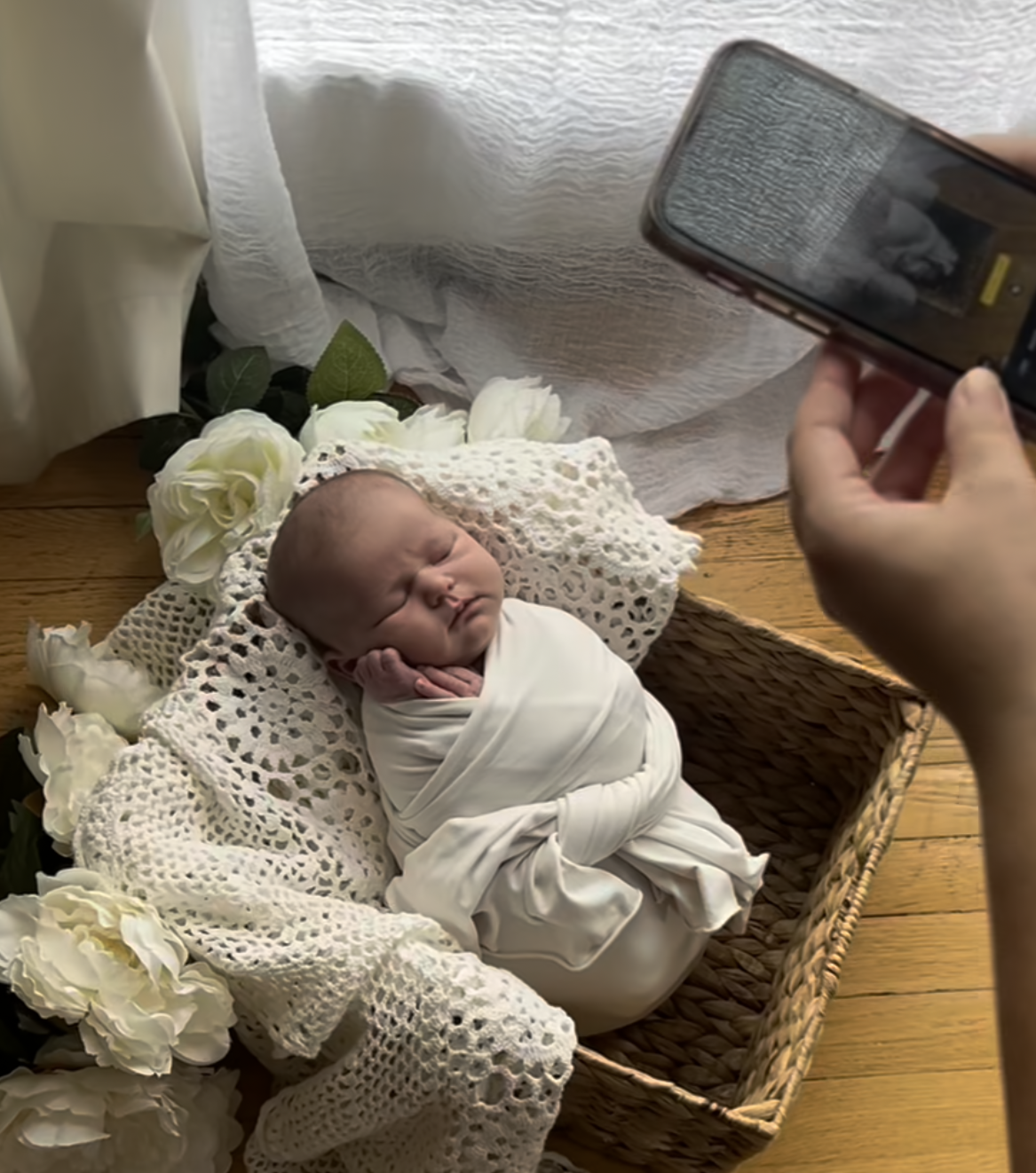Newborn baby swaddled in white blanket resting in wicker basket with white roses while someone photographs with smartphone.