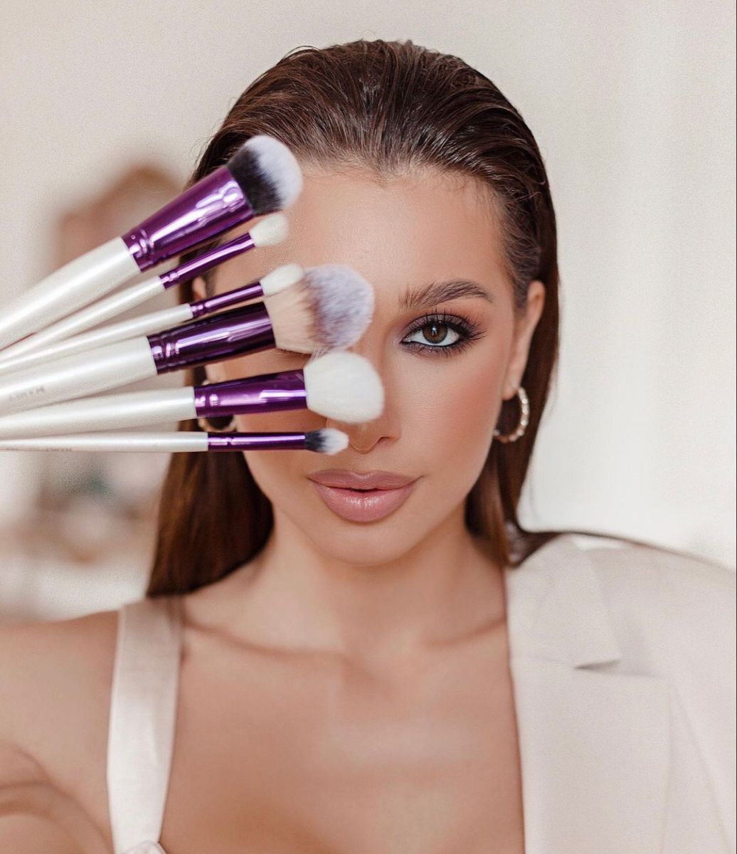 Woman holding purple and white makeup brushes fanned out in front of her face, wearing neutral makeup and hoop earrings.