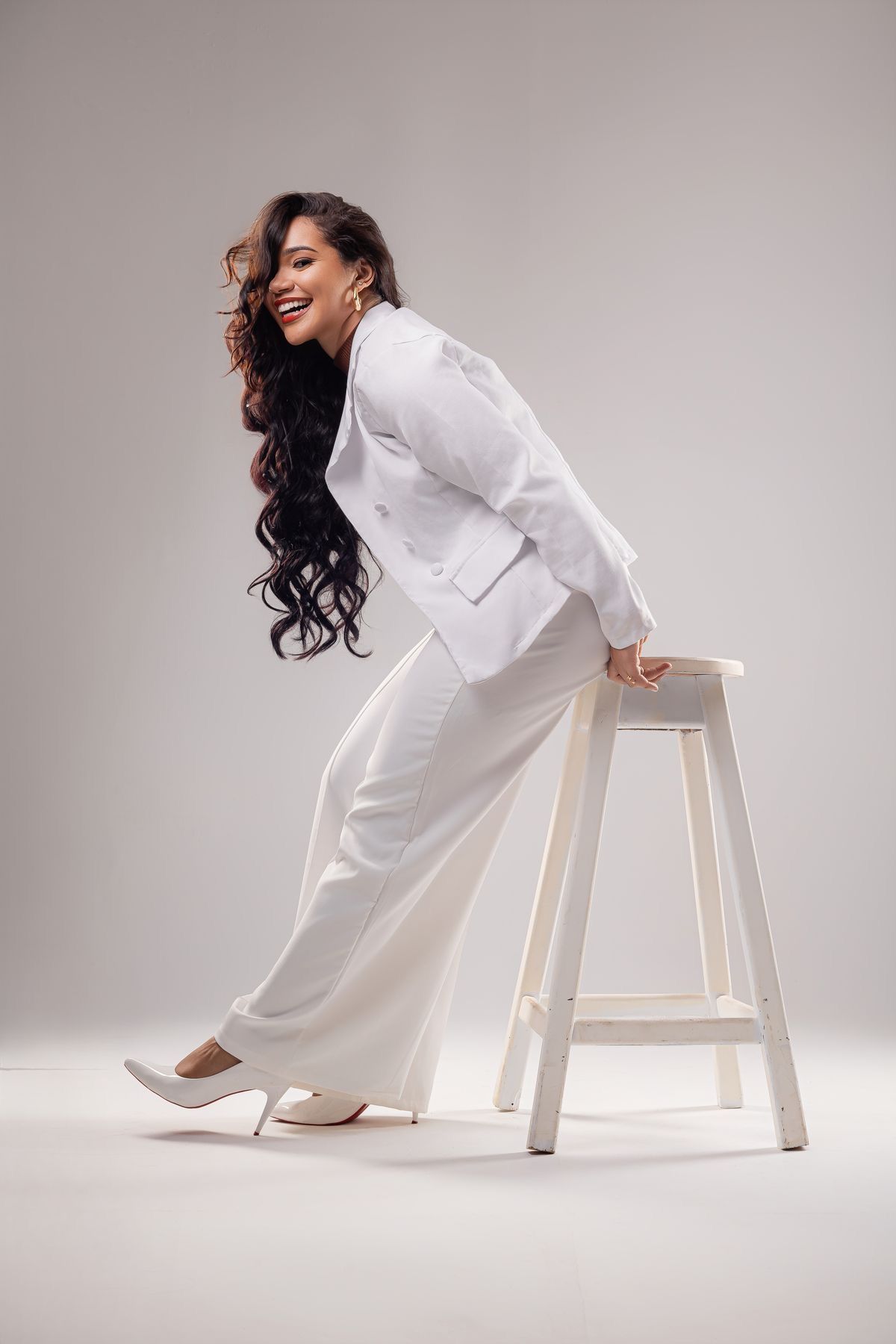 Smiling woman in white suit and heels leaning on a white stool against a light gray background.
