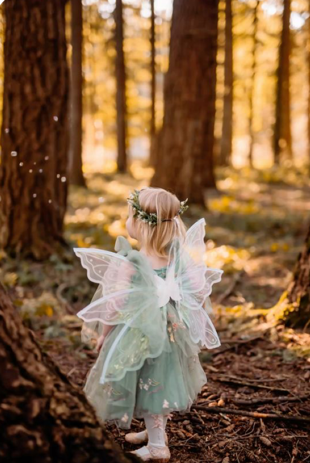 A young girl in a mint fairy costume with wings and floral crown stands in a sunlit forest.