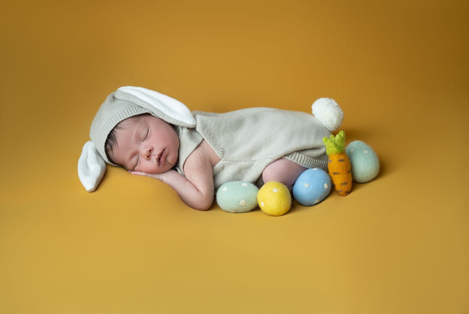 Newborn baby sleeping in bunny costume with Easter eggs and carrot toy on yellow background.