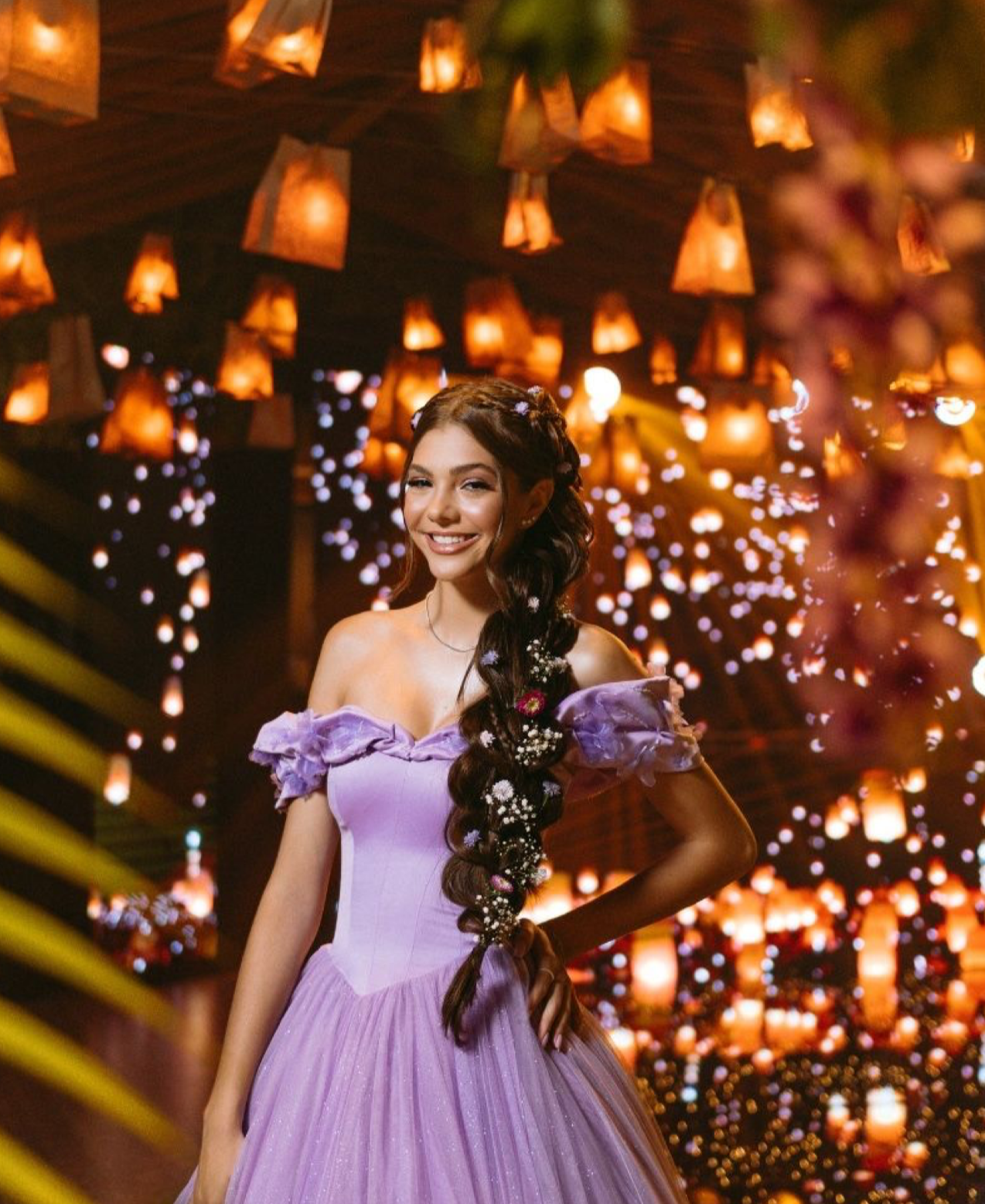 Young woman in lavender off-shoulder gown poses among glowing lanterns at an elegant quinceañera celebration.