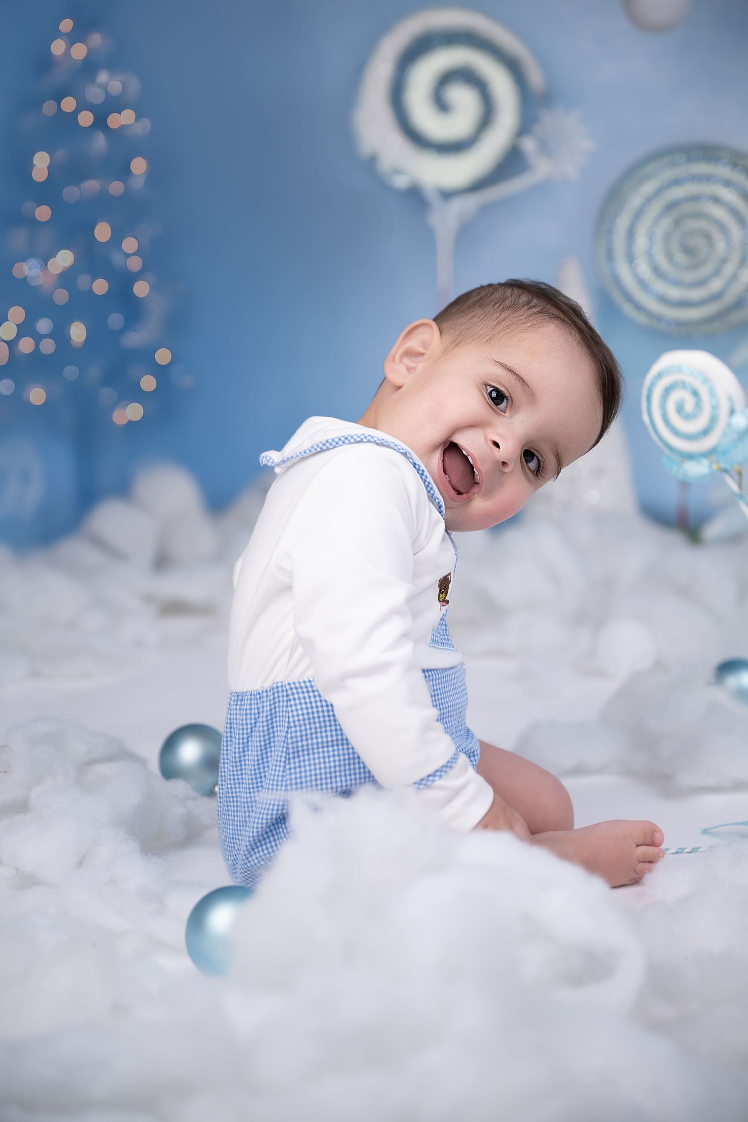 Happy baby in blue overalls sits among Christmas ornaments and candy decorations in a festive blue holiday setting.