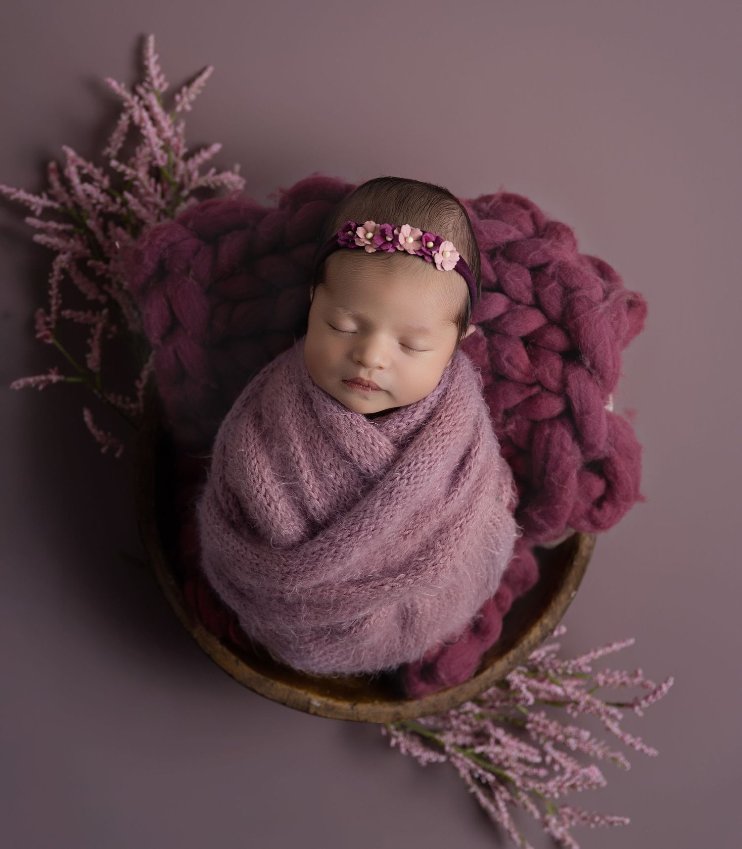 Newborn baby swaddled in purple wrap, sleeping in wooden bowl with floral headband and lavender sprigs.