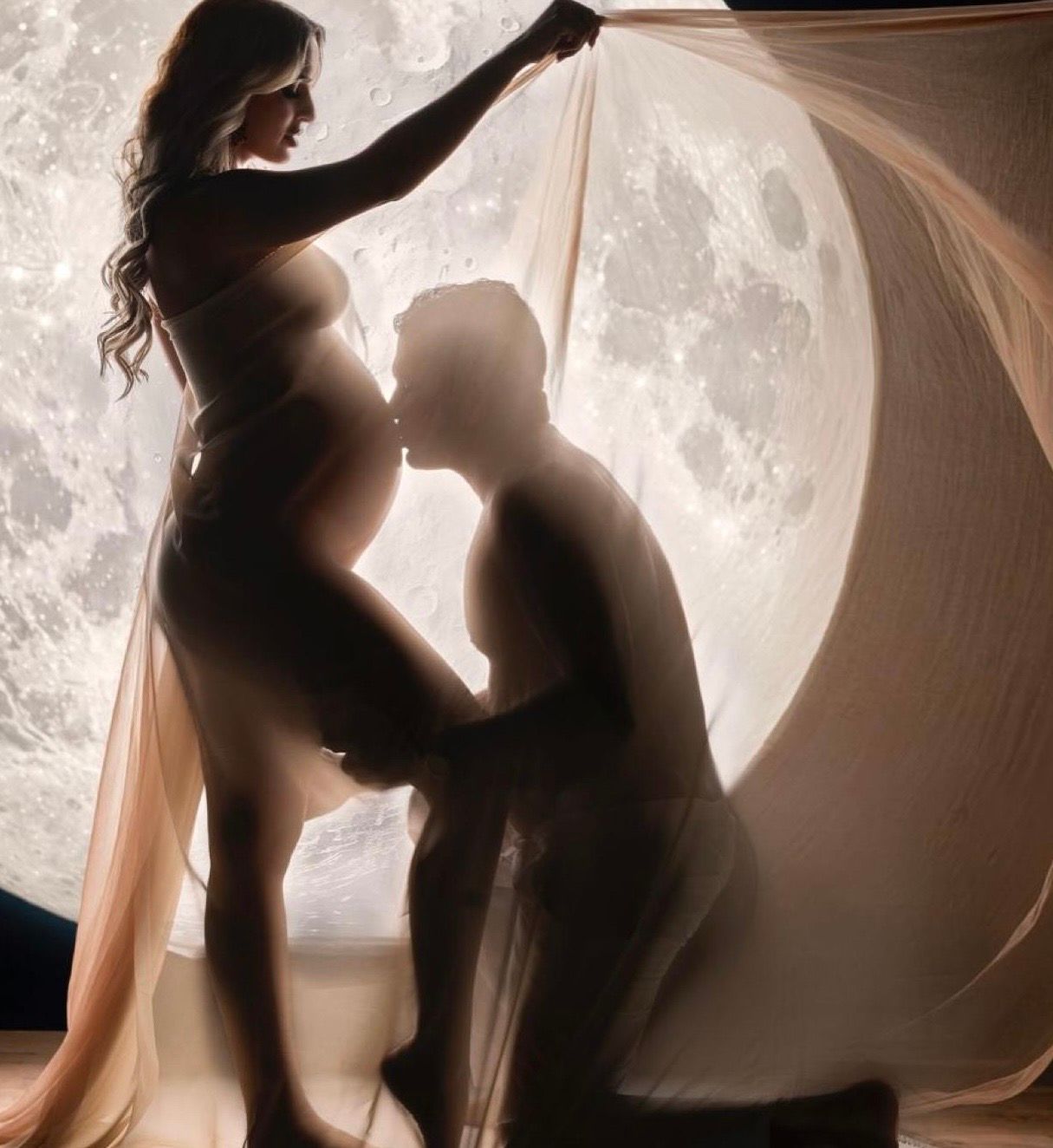 Romantic maternity silhouette of couple kissing in front of glowing moon backdrop with flowing sheer fabric.