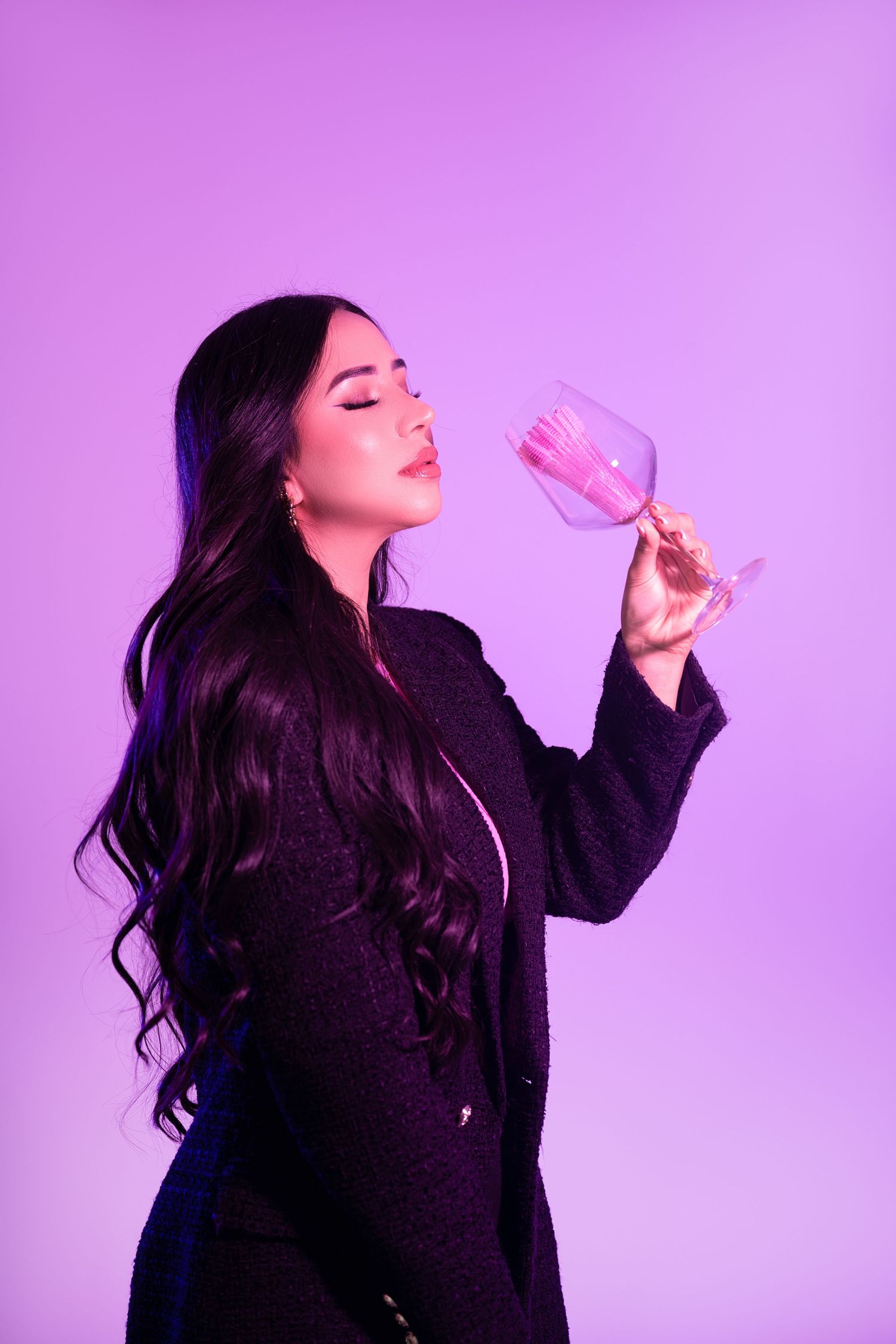 Fashion model in black outfit poses with pink wine glass against purple backdrop.