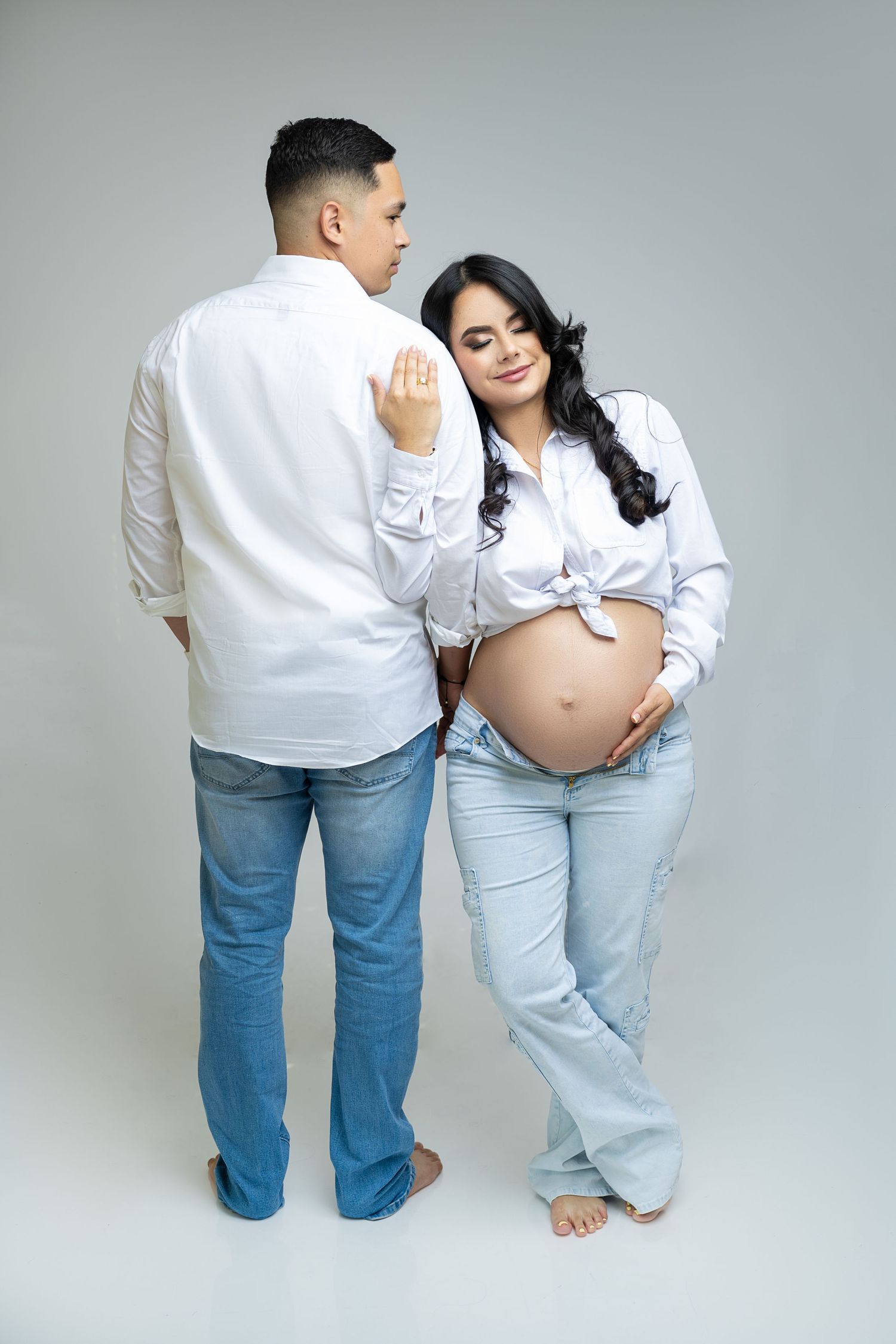 Couple wearing matching white shirts and jeans embrace during maternity photoshoot against gray backdrop.