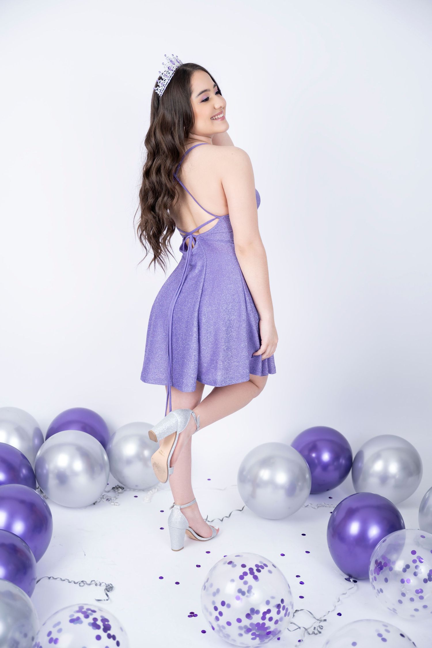 Playful pose in purple dress with silver and purple balloons on white background.