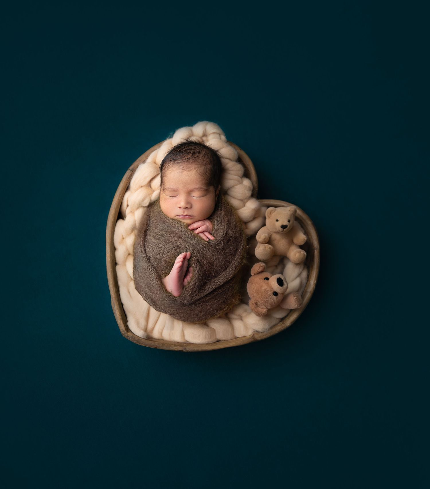 Newborn baby sleeping in heart-shaped basket with teddy bear on teal background.