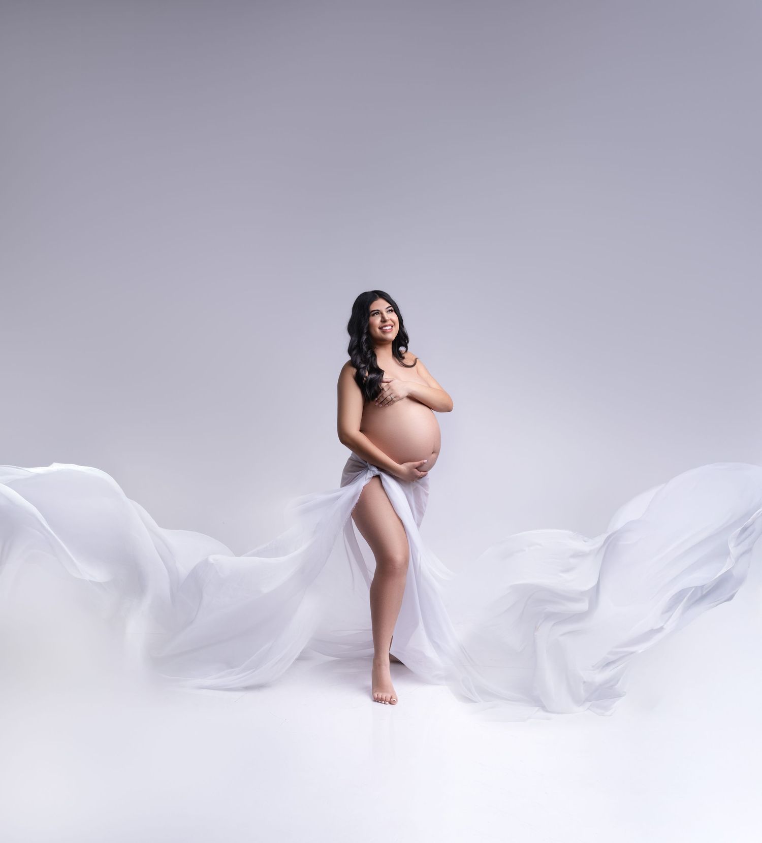 Artistic maternity photo with flowing white fabric against grey background.