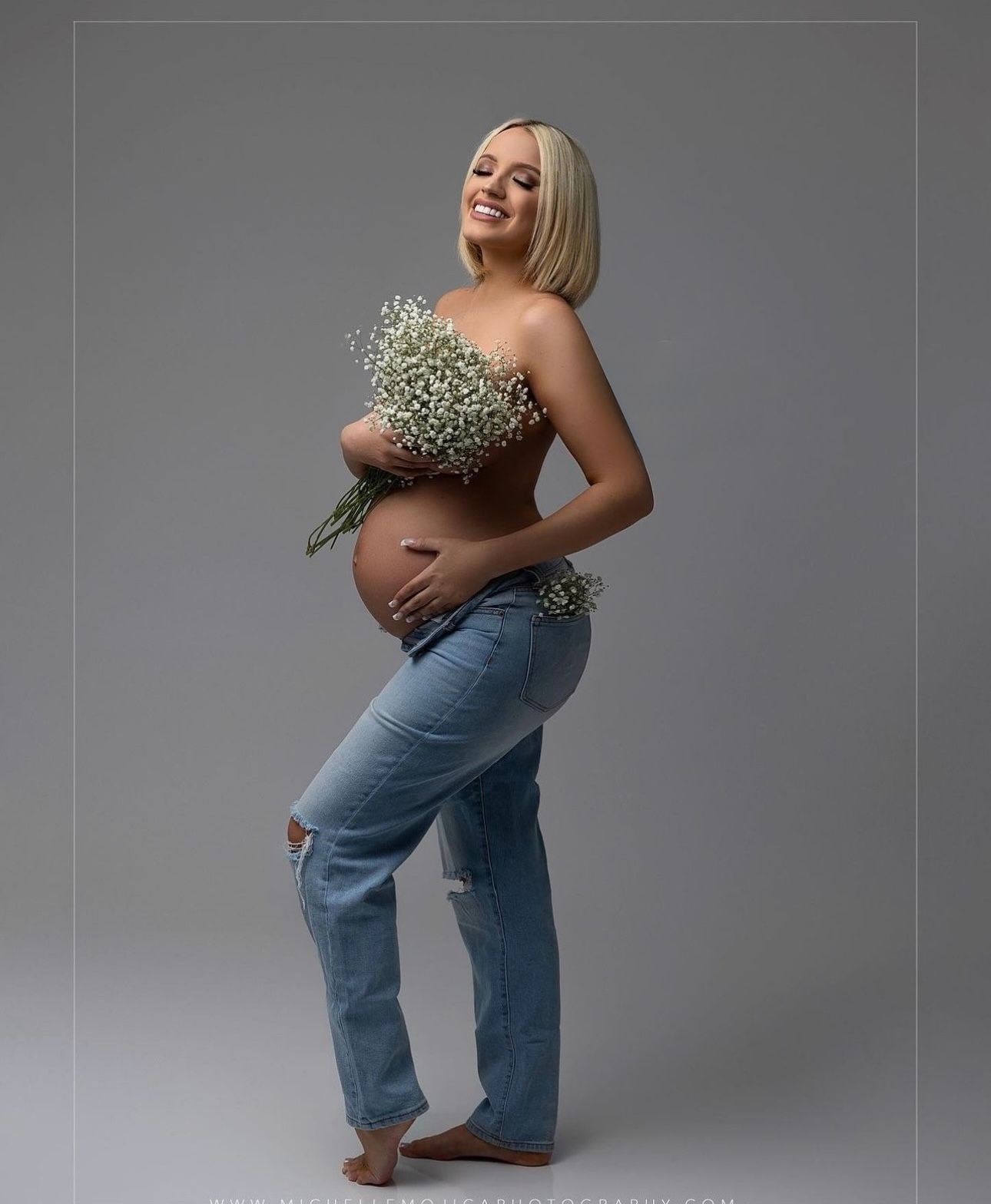 Pregnant woman in ripped jeans holding white baby's breath flowers, smiling in a maternity photoshoot against gray background.