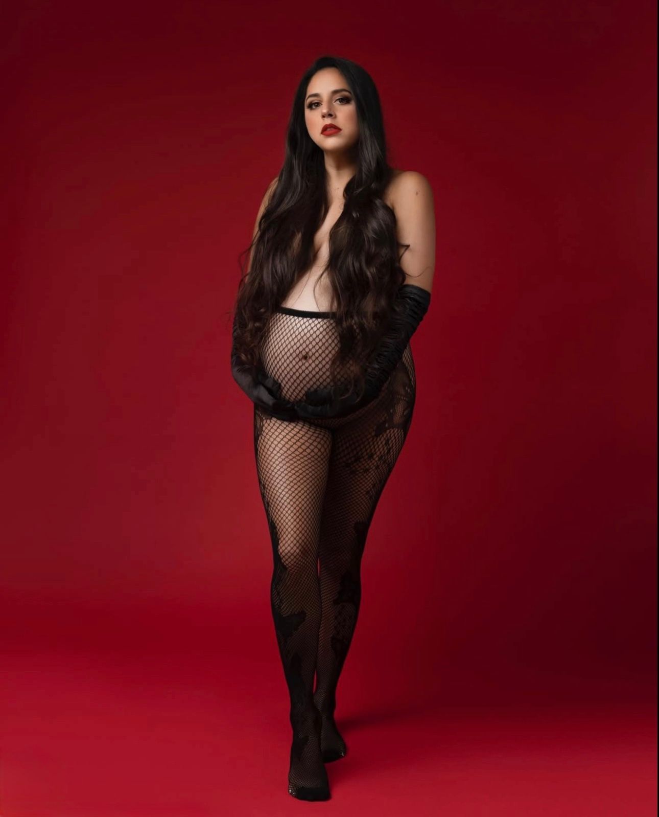 Woman in black fishnet bodysuit with long dark hair poses against red background.