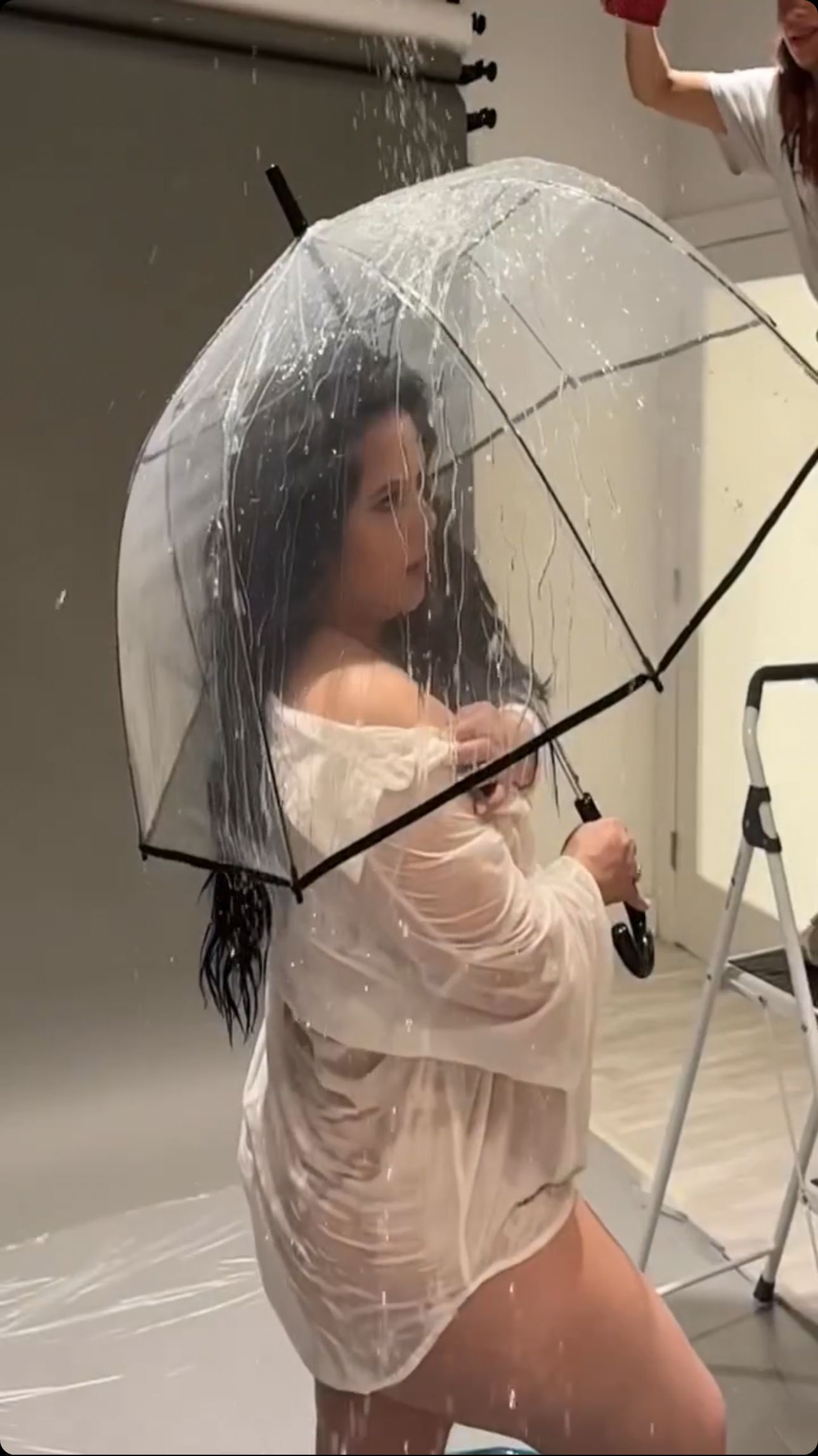 Woman holding clear bubble umbrella in wet white outfit during indoor rain photoshoot.