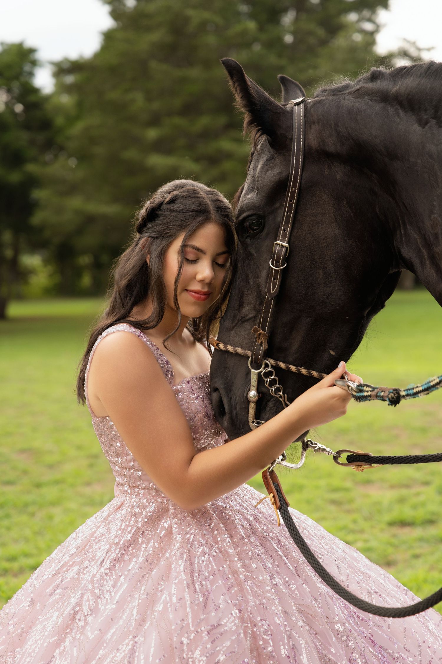 Young woman in pink sequin ball gown tenderly holds reins of black horse outdoors on green grass.