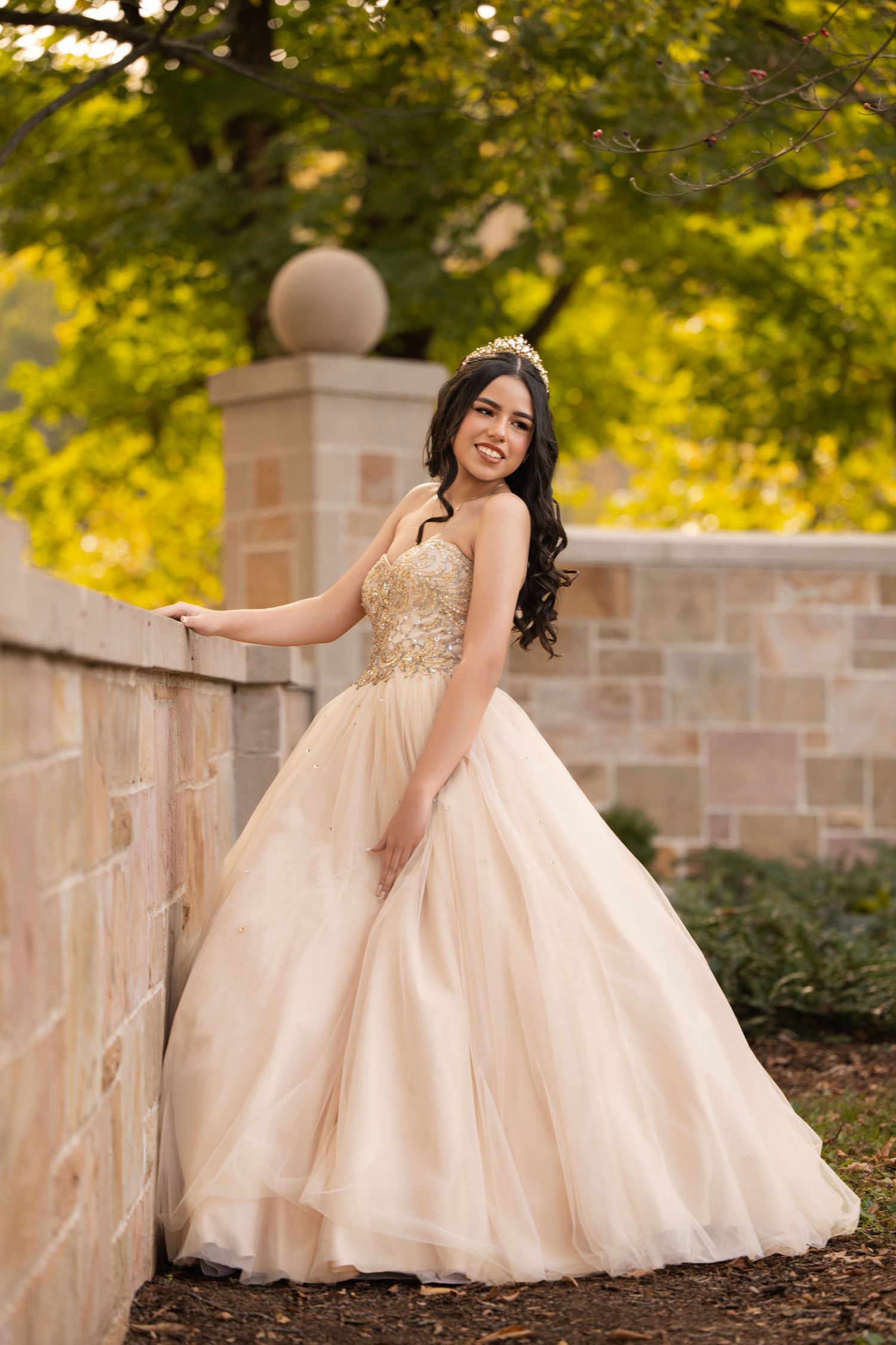 Young woman in gold quinceañera ballgown and tiara poses gracefully by stone wall in lush garden setting.
