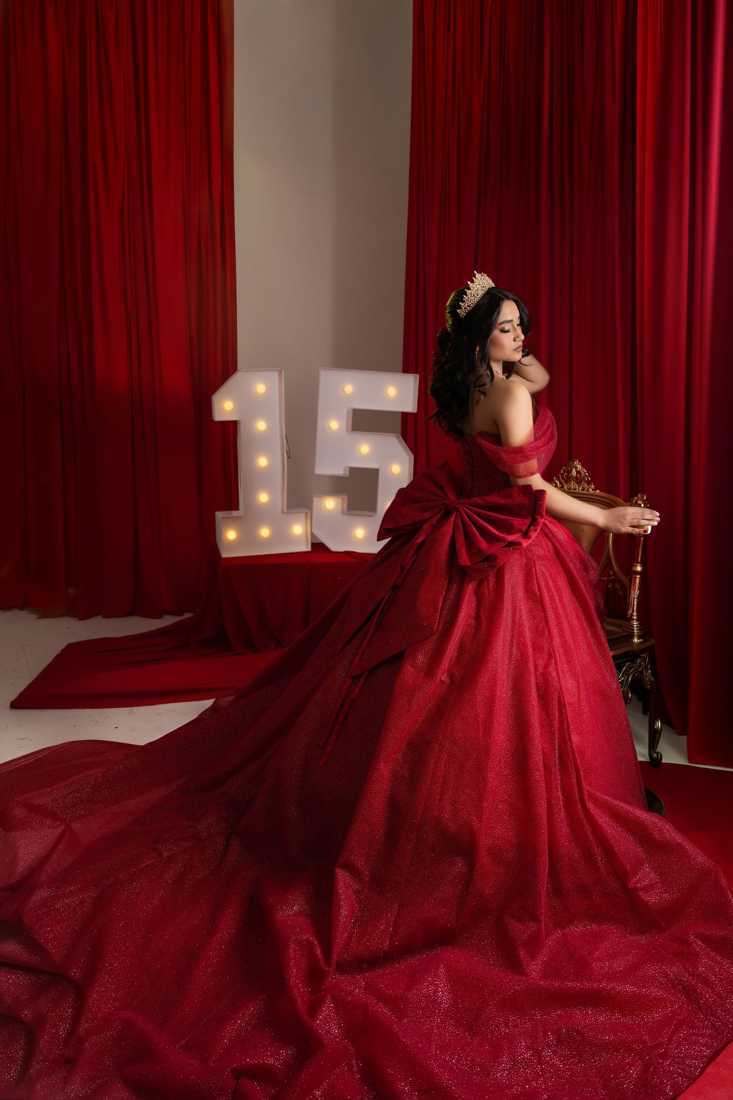 Young woman in elegant red ball gown with crown poses beside illuminated number 15 for quinceañera celebration.
