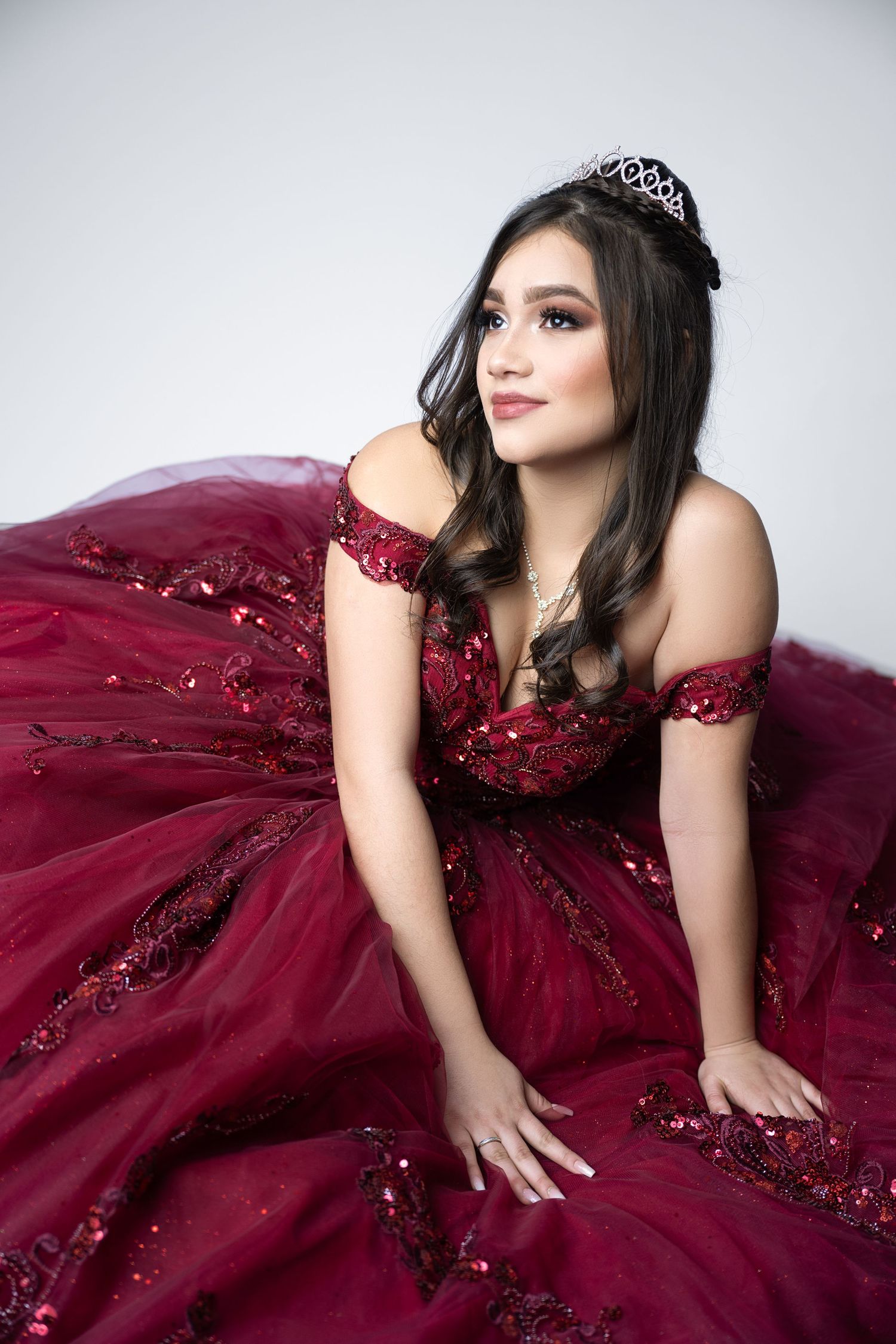Young woman in a red sequined ball gown and tiara poses gracefully for a quinceañera portrait.