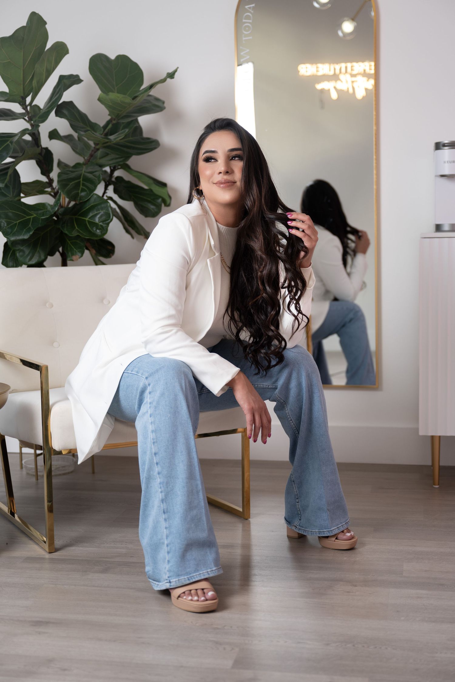 Woman in white blazer and flare jeans sits in modern salon chair, long curly hair, mirror reflection behind her.