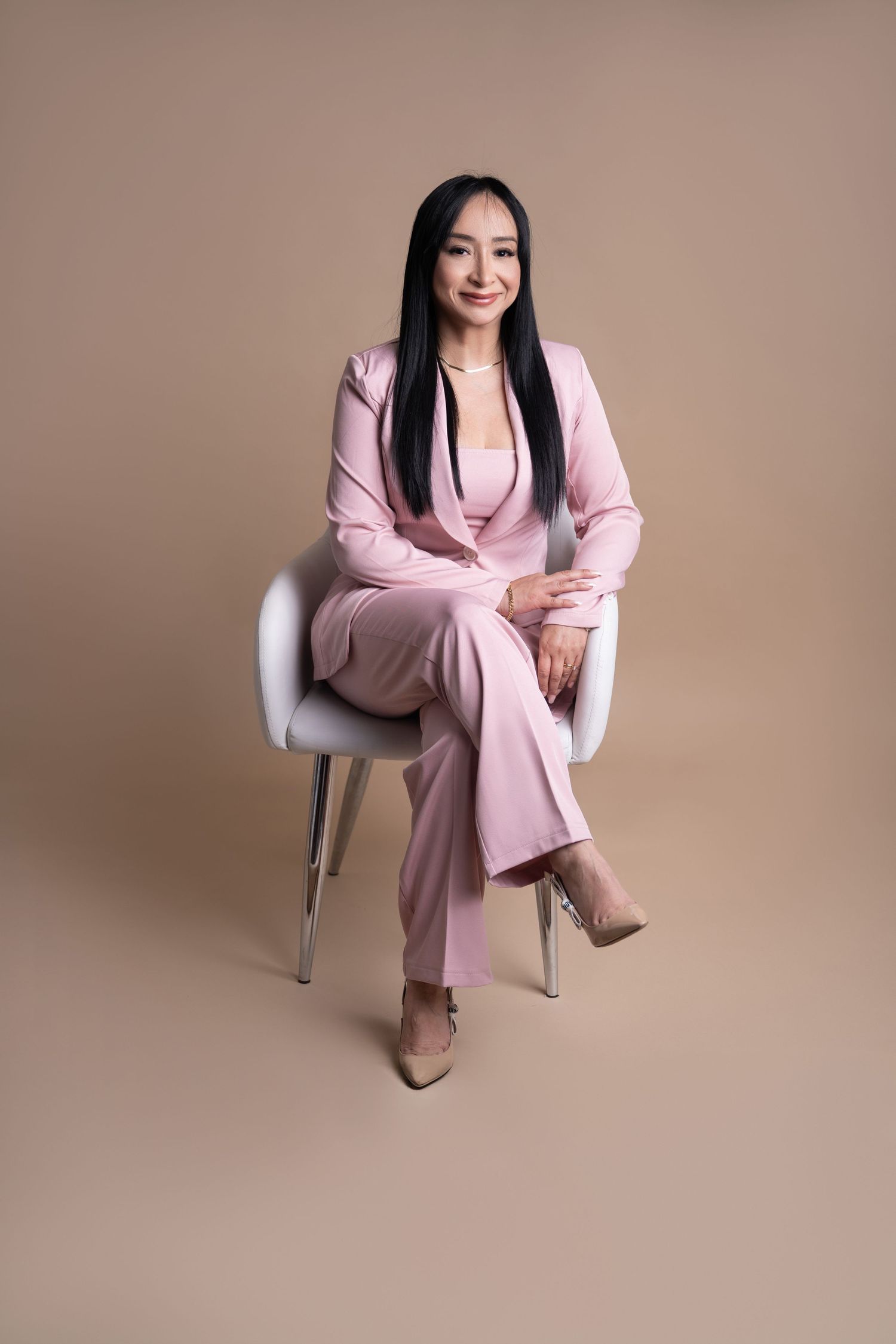 Woman in pink suit sitting confidently in white chair against beige background, professional portrait photo.
