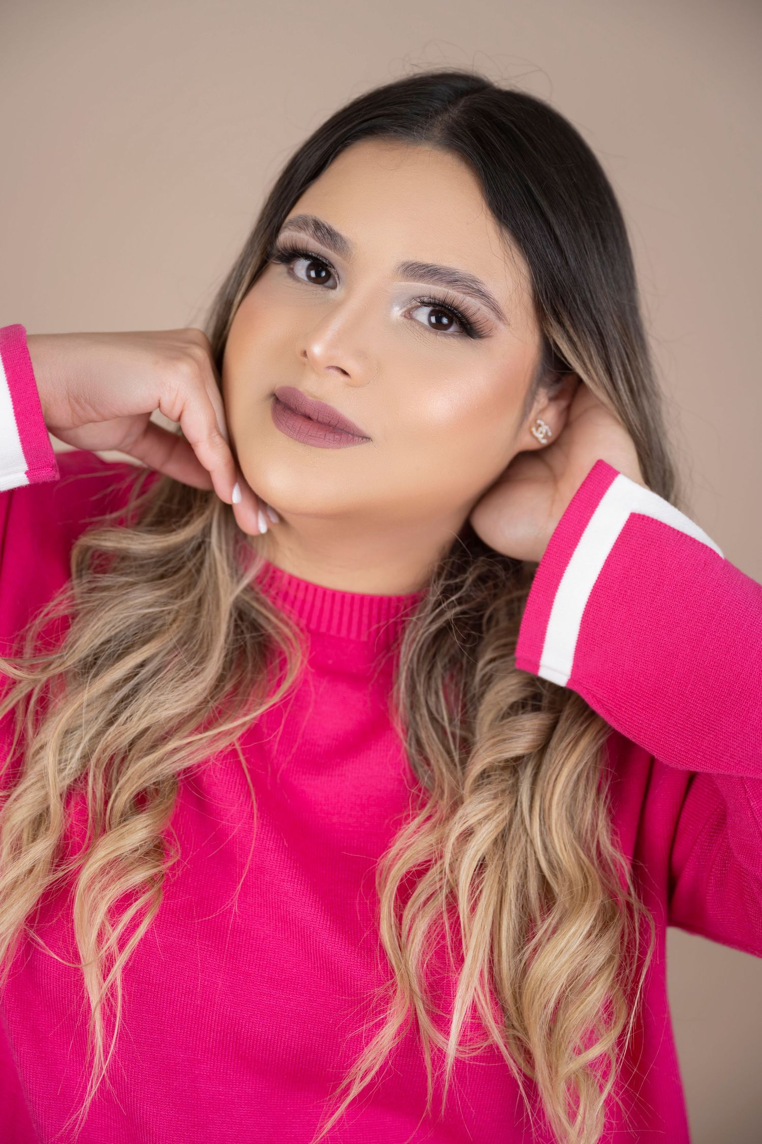 Woman with long wavy ombre hair and soft makeup poses with hands near her face, wearing a bright pink sweater against a beige backdrop.
