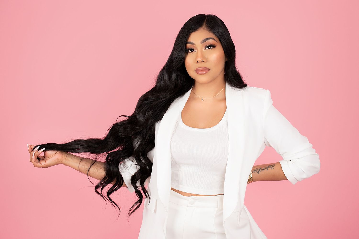 Woman with long, wavy black hair holds a strand while posing in a white blazer and matching crop top against a bright pink background.