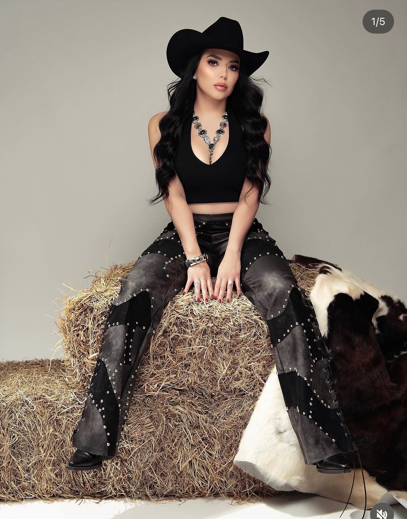 Woman in black cowboy hat, crop top, and studded flare pants sitting on hay bale with cowhide in western fashion shoot.